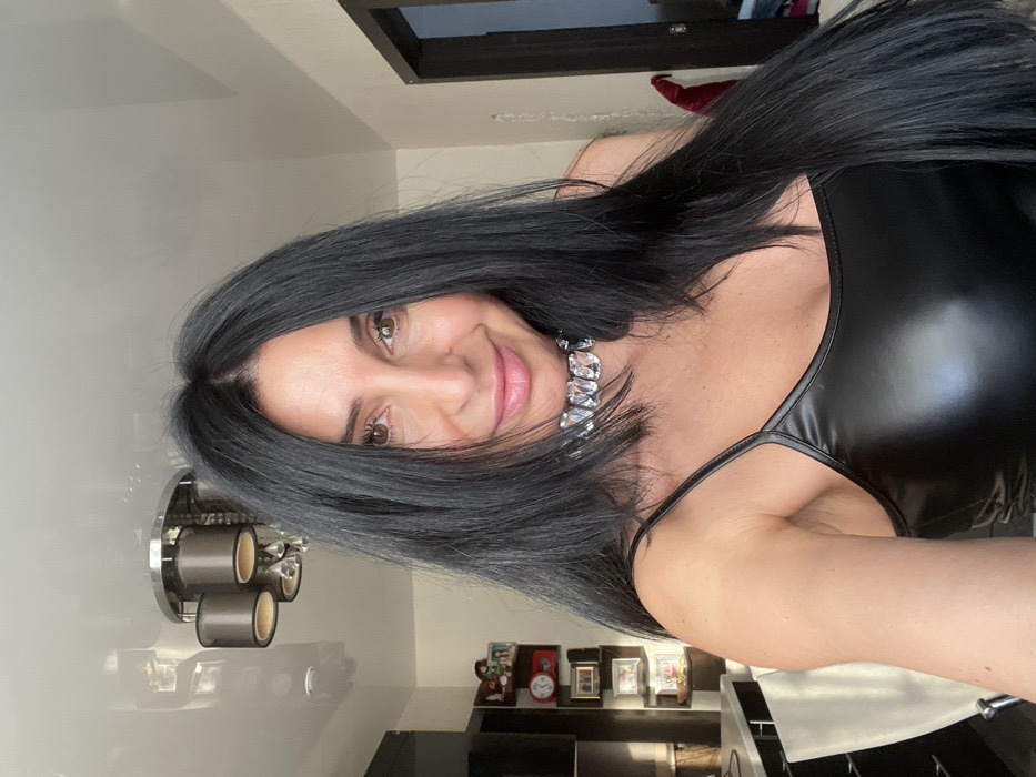 The image features a woman posing indoors, looking directly at the camera with a friendly, closed-mouth smile. She has long, straight, dark (likely black or very dark brown) hair, parted on the side, which falls over her shoulders and partially across her face. Her complexion is fair, and she has brown eyes and well-defined eyebrows. Her lips are a light pink shade.
She is wearing a form-fitting, shiny black top, possibly made of faux leather or a similar material, with thin spaghetti straps visible on her right shoulder. Around her neck, she wears a prominent, chunky silver-toned necklace or choker adorned with large, clear, rectangular or cushion-cut gemstones or crystals.
The background suggests an indoor setting, likely a home. To the left, a modern light fixture with three cylindrical, dark-colored shades is mounted on a light-colored wall. Below this, a dark-colored shelf or surface holds several small framed photos or decorative items and a dark mug. In the upper right corner, a portion of a dark-framed object, possibly a window or a piece of furniture, is visible. The lighting in the scene appears bright and even, indicating a well-lit room. No discernible text is visible in the image.