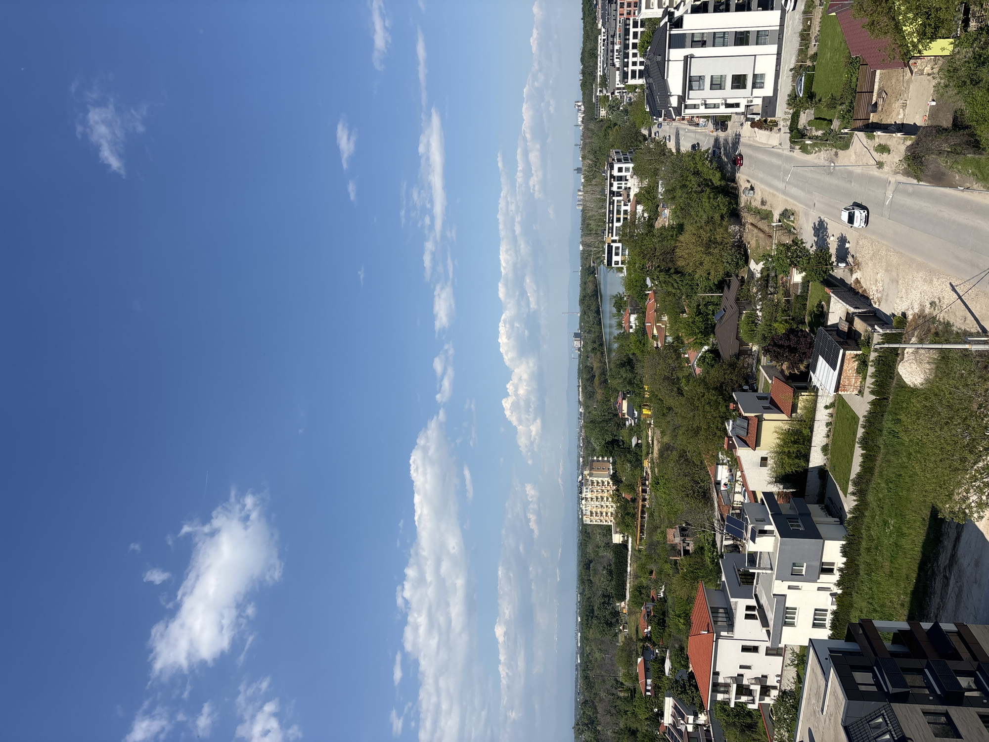 The image presents a view from an elevated position, possibly a high-rise building or a hill, overlooking a neighborhood in Plovdiv, Bulgaria. The sky takes up a significant portion of the frame, displaying a clear blue gradient with scattered cumulus clouds. The lower part of the image captures a mix of residential and green areas. Modern, multi-story apartment buildings are interspersed with older houses, most of which have red tile roofs. Trees and landscaping are abundant, partially obscuring the buildings. A road runs through the scene, with one white car driving along it. In the distance, a body of water, possibly the Maritsa River, is visible. No people are clearly discernible in the image.