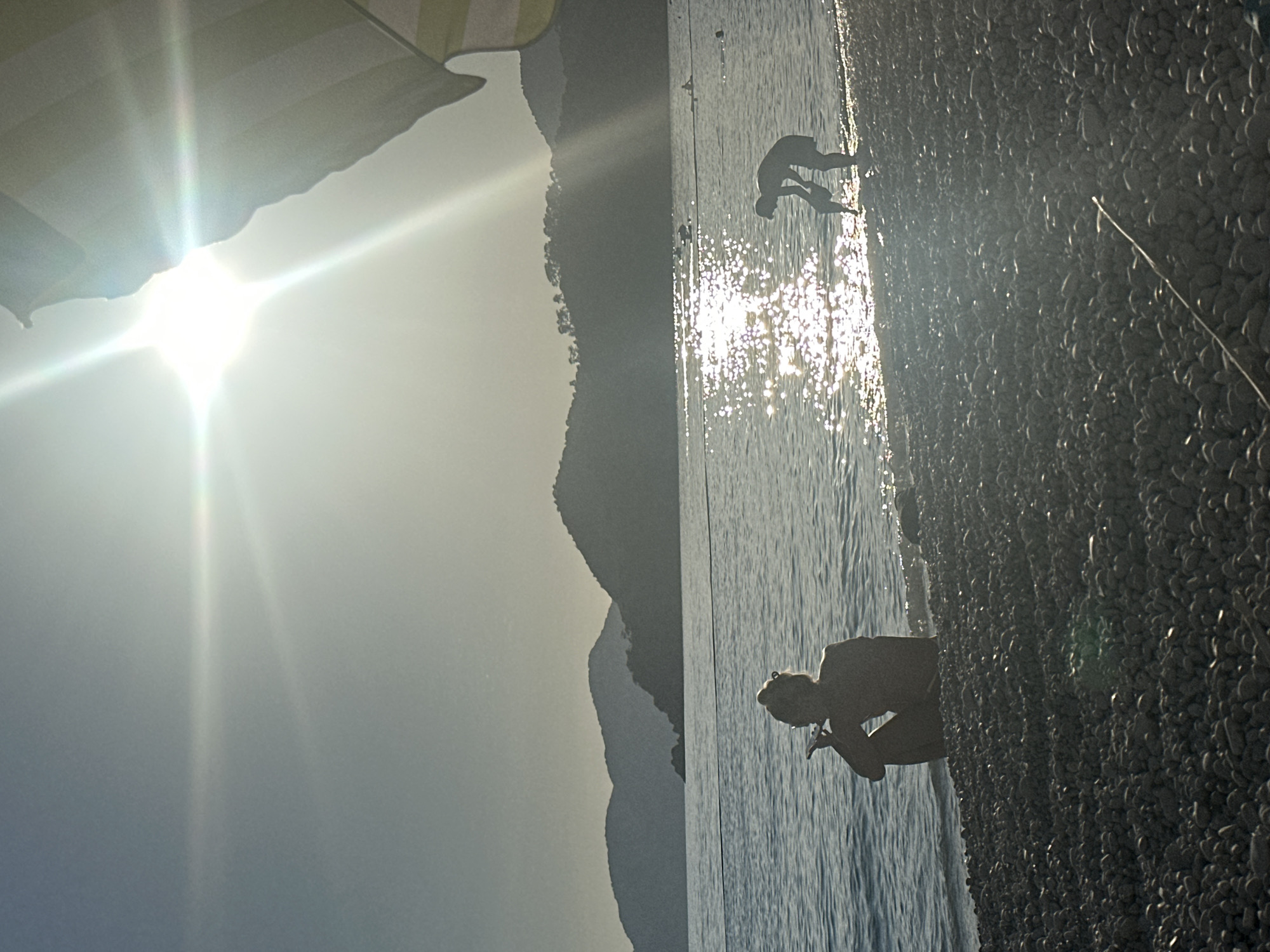 This image depicts a bright, sunny day at a pebble beach, likely in Fethiye, Turkey, given the context. The scene is dominated by a powerful sun flare emanating from the upper left, creating a strong, almost ethereal glow across the sky and reflecting intensely off the water. Part of a striped beach umbrella or canopy is visible in the upper left corner, partially obscuring the direct sunlight.
In the mid-ground, a range of dark, rugged mountains stretches horizontally, silhouetted against the bright, hazy sky. To the right, the calm sea shimmers with countless points of light from the sun's reflection, suggesting clear waters.
Two individuals are silhouetted against the sparkling water. In the mid-ground on the right, a person stands or wades in the shallow water, facing away from the viewer. Closer to the foreground, towards the bottom right, another person is seen crouched or bending over at the water's edge, their form also a dark silhouette against the bright reflections. The foreground consists of a coarse pebble beach, and a thin, white line, possibly a fishing line or string, extends from the lower right corner onto the pebbles.
The time of day appears to be mid-day or late afternoon, characterized by the strong, direct sunlight and pronounced lens flare. The weather is clear and sunny. The overall impression is one of a peaceful, leisurely day at a beautiful coastal location. No visible text is present in the image.