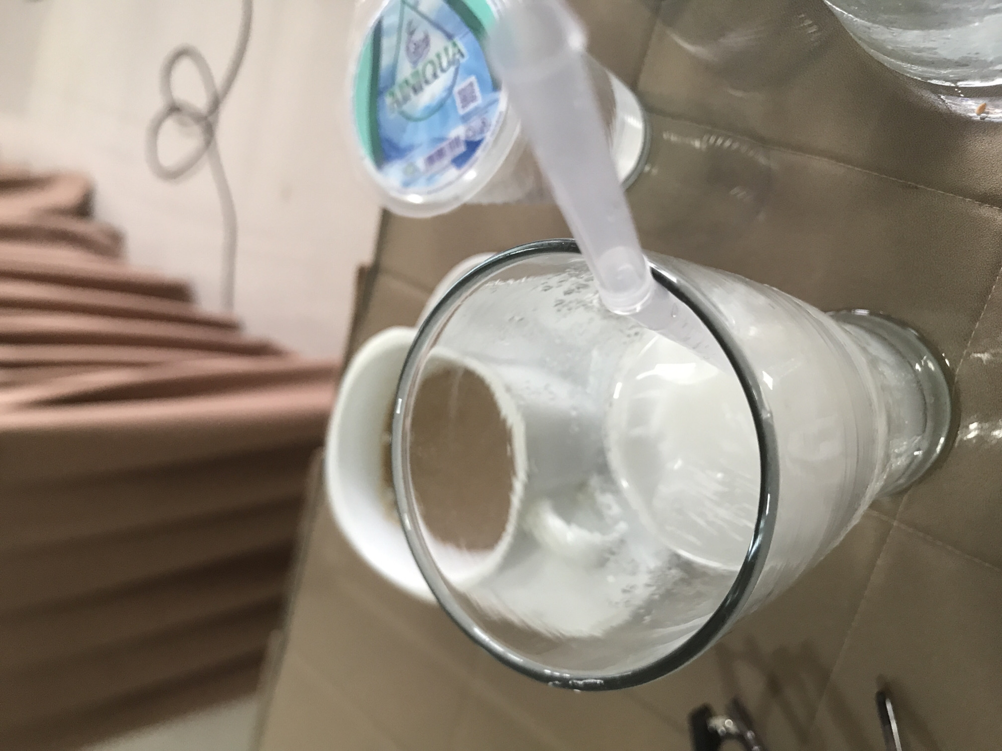 An indoor scene shows a close-up, eye-level shot of a table with drinks. In the foreground, a clear glass filled with a white beverage, likely milk or a smoothie, is positioned on the right side of the frame. A translucent white straw extends from the glass. To the left of this glass, a white mug filled with a brown liquid, presumably coffee, is visible. Further to the right, a small plastic cup with a blue and white label, possibly bottled water, is present. The background features a blurred view of a brown curtain. The table surface appears to be a textured, brown material. The overall setting suggests a casual dining or beverage service environment. The image is not sufficient to determine the specific city of Lhokseumawe, Indonesia.