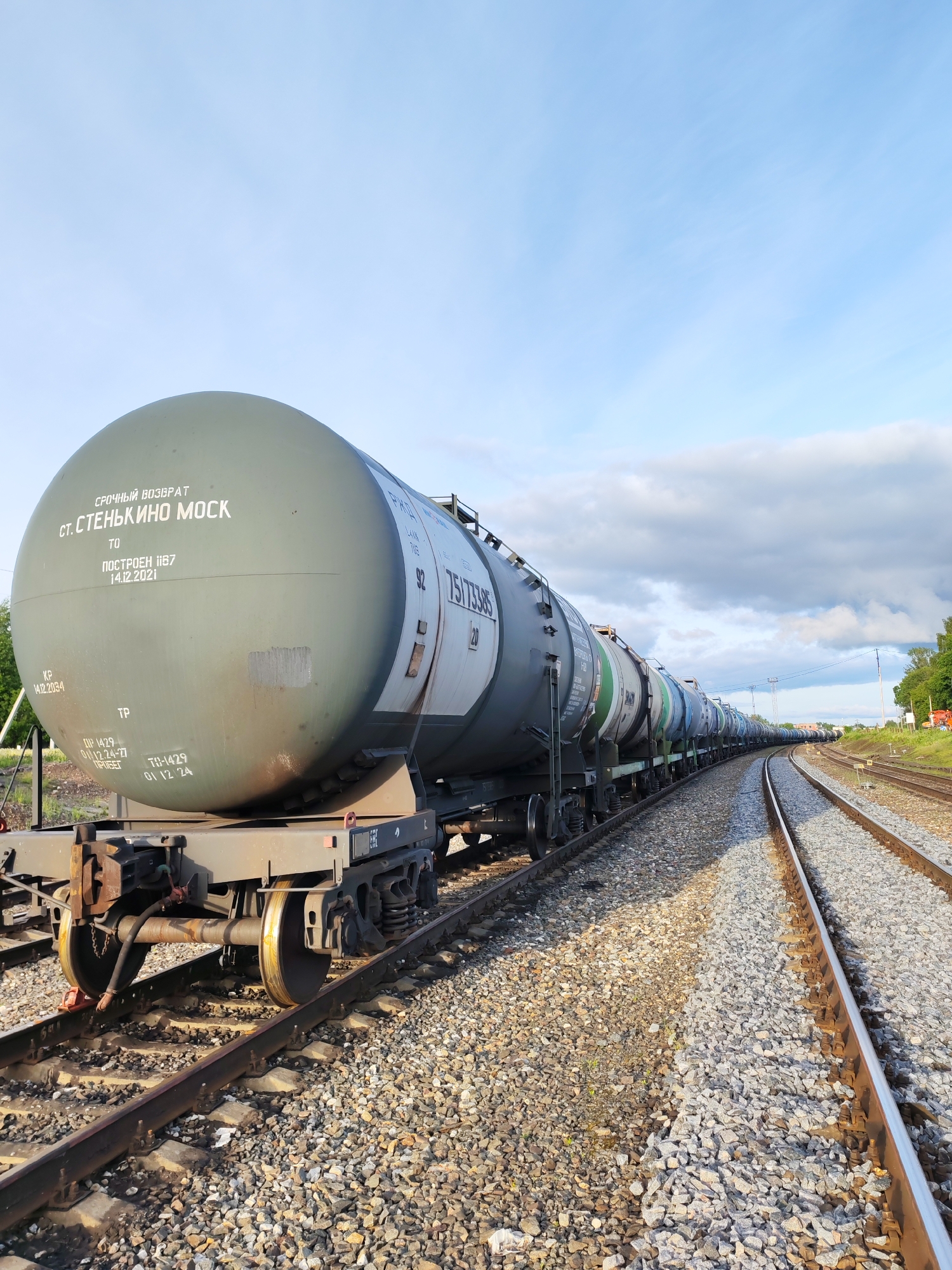 The image depicts a long line of tanker railcars parked on a railroad track, curving into the distance on a clear day with a blue sky and scattered white clouds. The tanks are gray and cylindrical with domed ends, with Russian text visible on the first tank car. Text says: URGENT RETURN. The location is specified as Bol'shie Mkhi, Russia. The foreground shows the ballast of the railbed consisting of gray gravel. A second track runs parallel to the first. Vegetation and what appears to be a building can be seen in the background to the right. No people are visible.