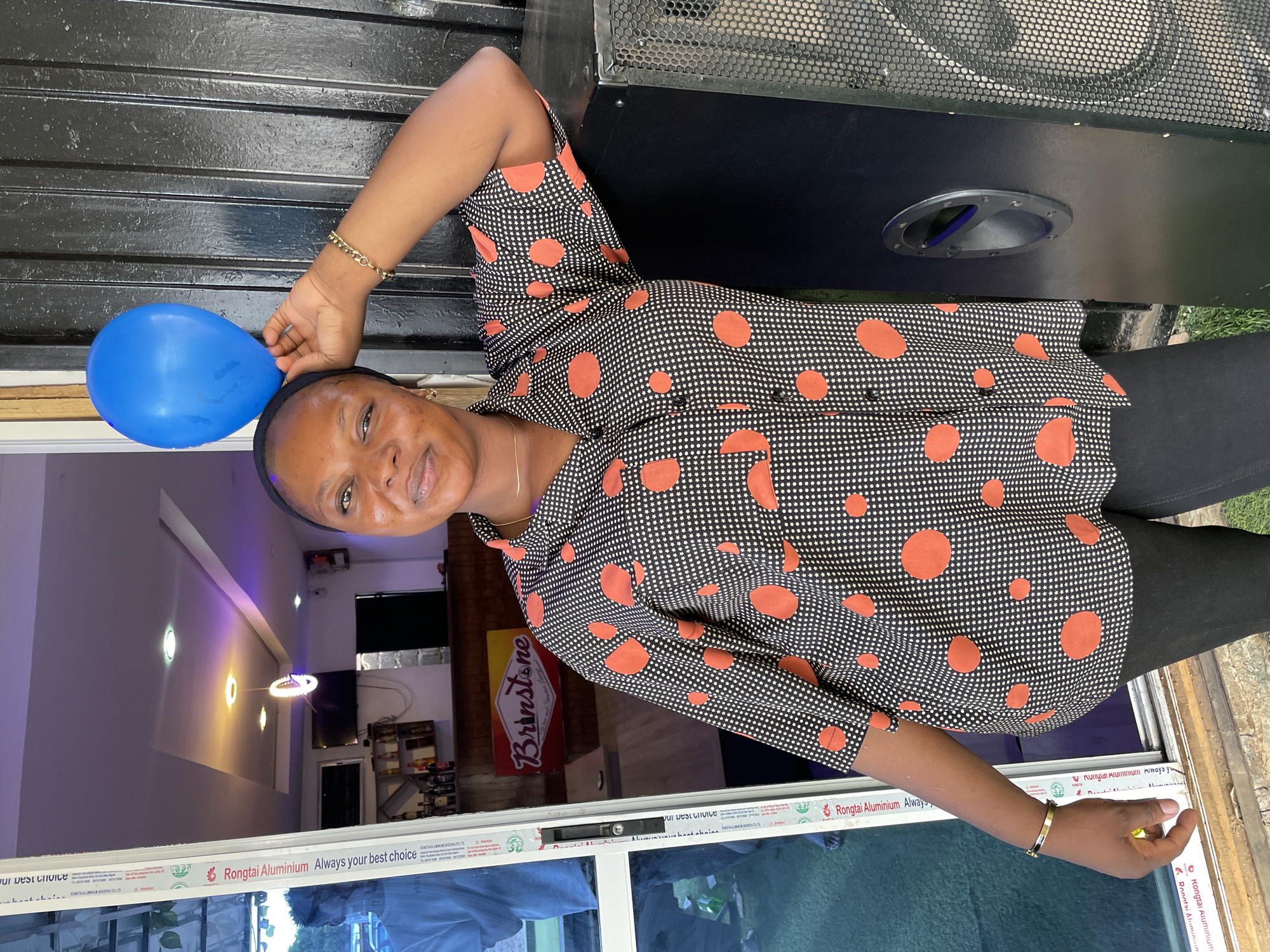 A woman is holding a blue balloon. She is wearing a black and white polka-dot shirt with red spots, black pants, and jewelry on her wrist. She is standing indoors near a window with the text "Rongtai Aluminium Always your best choice". Behind her is a sign that reads "Brinstone". There is a speaker box next to her and the scene appears to be located in Jos, Nigeria.
