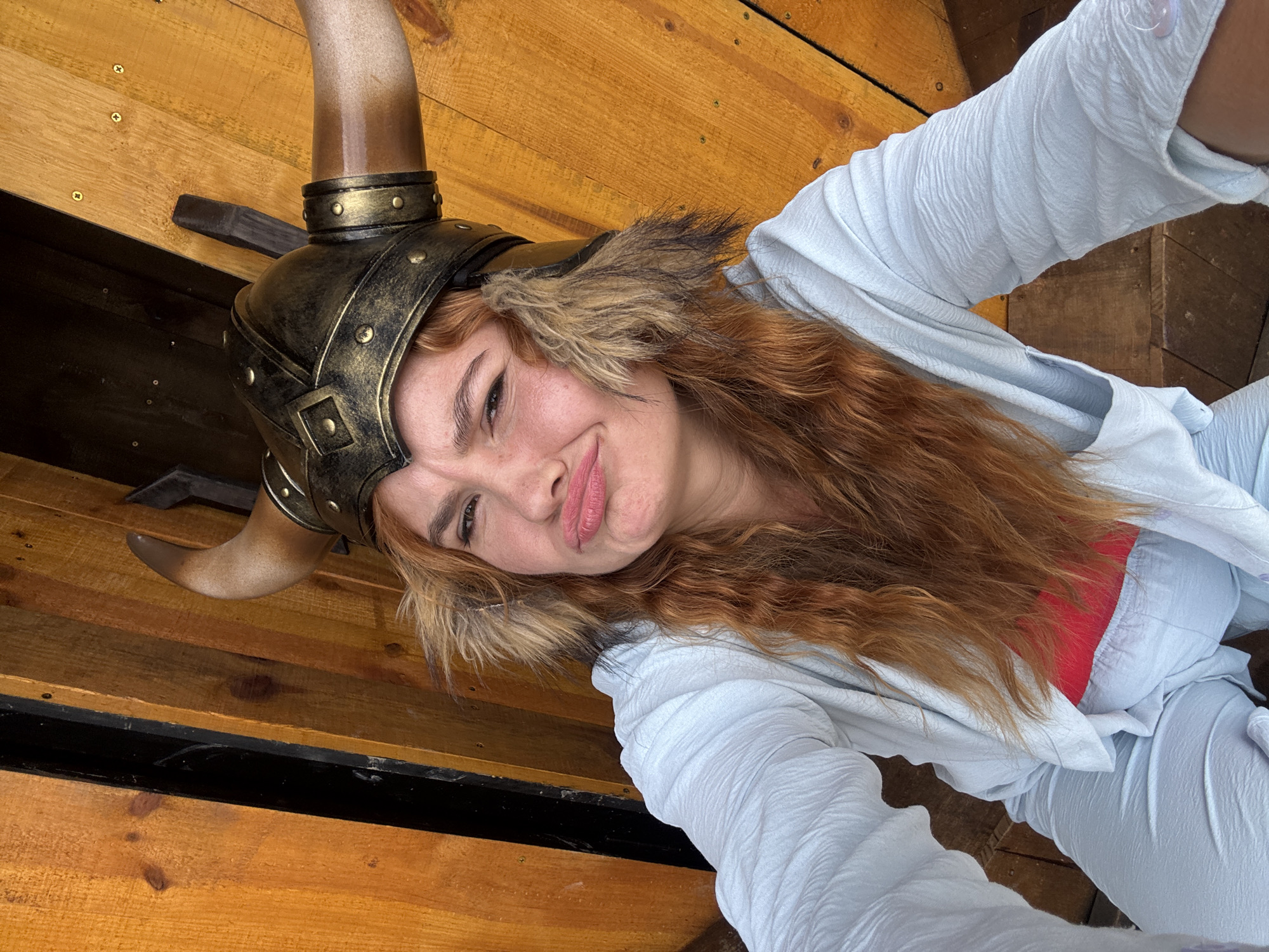 A woman with long, wavy red hair is wearing a Viking helmet with horns and fur trim. She is also wearing a light blue outfit and a red top. She is making a puckered face at the camera. The background appears to be made of wooden planks, possibly part of a boat or structure. The location is not specified in the city of Thôn 1, Vietnam.