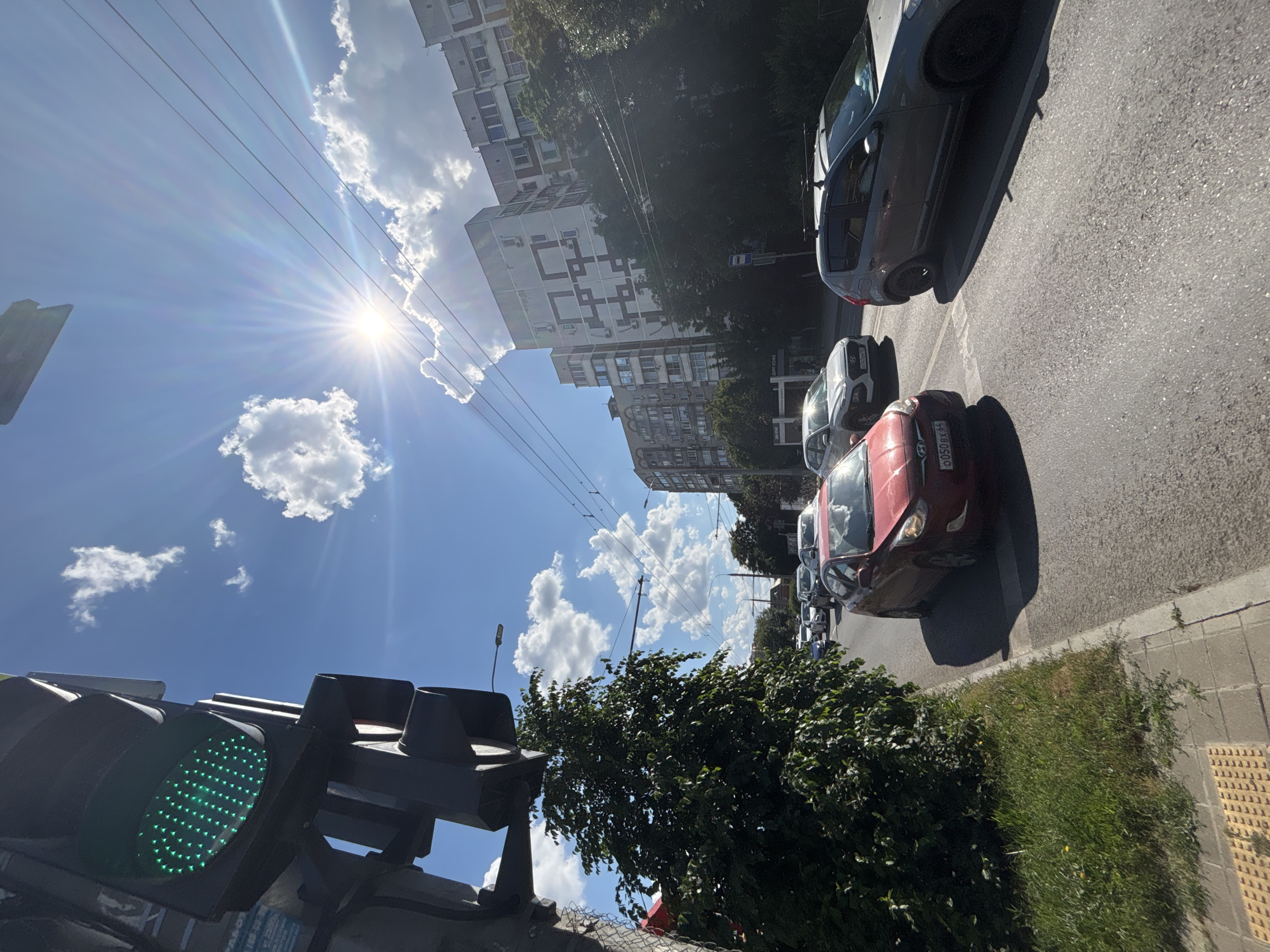 This is a vertical, eye-level, exterior shot taken on a sunny day in Krasnodar, Russia. The image features a street scene with several cars parked or driving on the right side of the road, as seen from the perspective of someone facing a traffic light. The sky is bright blue with scattered white clouds, and the sun is shining intensely, creating lens flare.
On the left, a traffic light is visible, with its green light illuminated. In the background, tall apartment buildings with multiple windows line the street, interspersed with trees that are full of green leaves, suggesting a warm season. The buildings appear to be residential, with balconies and various architectural details.
The cars visible include a red car in the foreground with a license plate that reads "6050PY 54", a silver car, and other vehicles further down the street. Power lines crisscross the sky. The overall atmosphere is bright and active, indicative of a typical day in a city. No people are clearly visible in the image. The ground is paved asphalt with a sidewalk and grass area on the right. The time of day appears to be late morning or early afternoon due to the bright sunlight and the angle of the shadows.