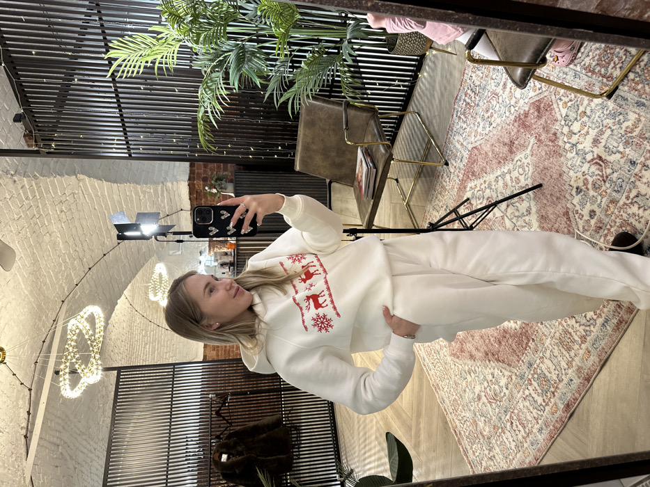 A blonde woman in a white reindeer-themed sweatsuit takes a mirror selfie. Her phone is held up to the upper right. The reflection captures a room with a vaulted ceiling, modern lighting, and a metal-slatted wall behind the woman. There's a green plant in the bottom right and various pieces of furniture behind her, including a chair with books. A patterned rug is seen on the right-hand side of the frame.