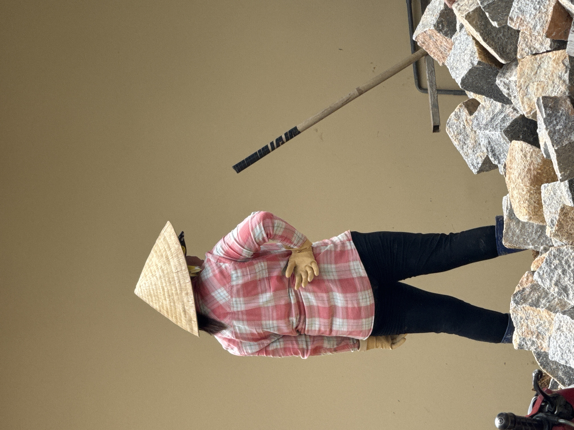 In Da Lat, Vietnam, a person wearing a traditional conical hat stands with their back to the camera. They are dressed in a pink and white plaid shirt, black pants, and blue boots. The person has their left hand on their hip and is wearing yellow gloves. In the background, a stack of rough-hewn granite stones is visible, with a tool, possibly a rake or shovel, leaning against them. The scene is set against a plain, neutral-colored wall.