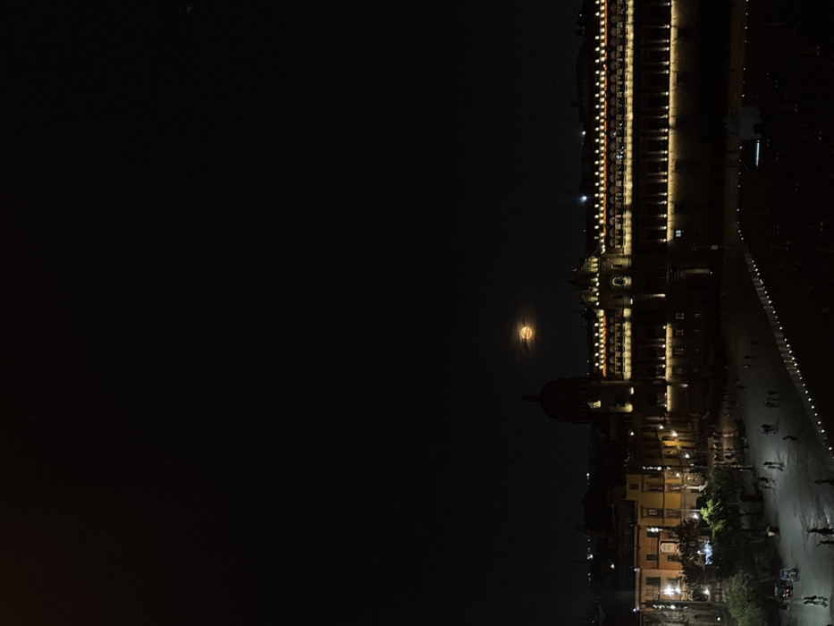 A nighttime cityscape with buildings illuminated along a waterfront. A full moon is visible above a domed structure. Lights line the buildings facing the water, reflecting in the dark surface. People are seen walking along the embankment. The sky is predominantly black. The image is captured in a dark, moody tone.