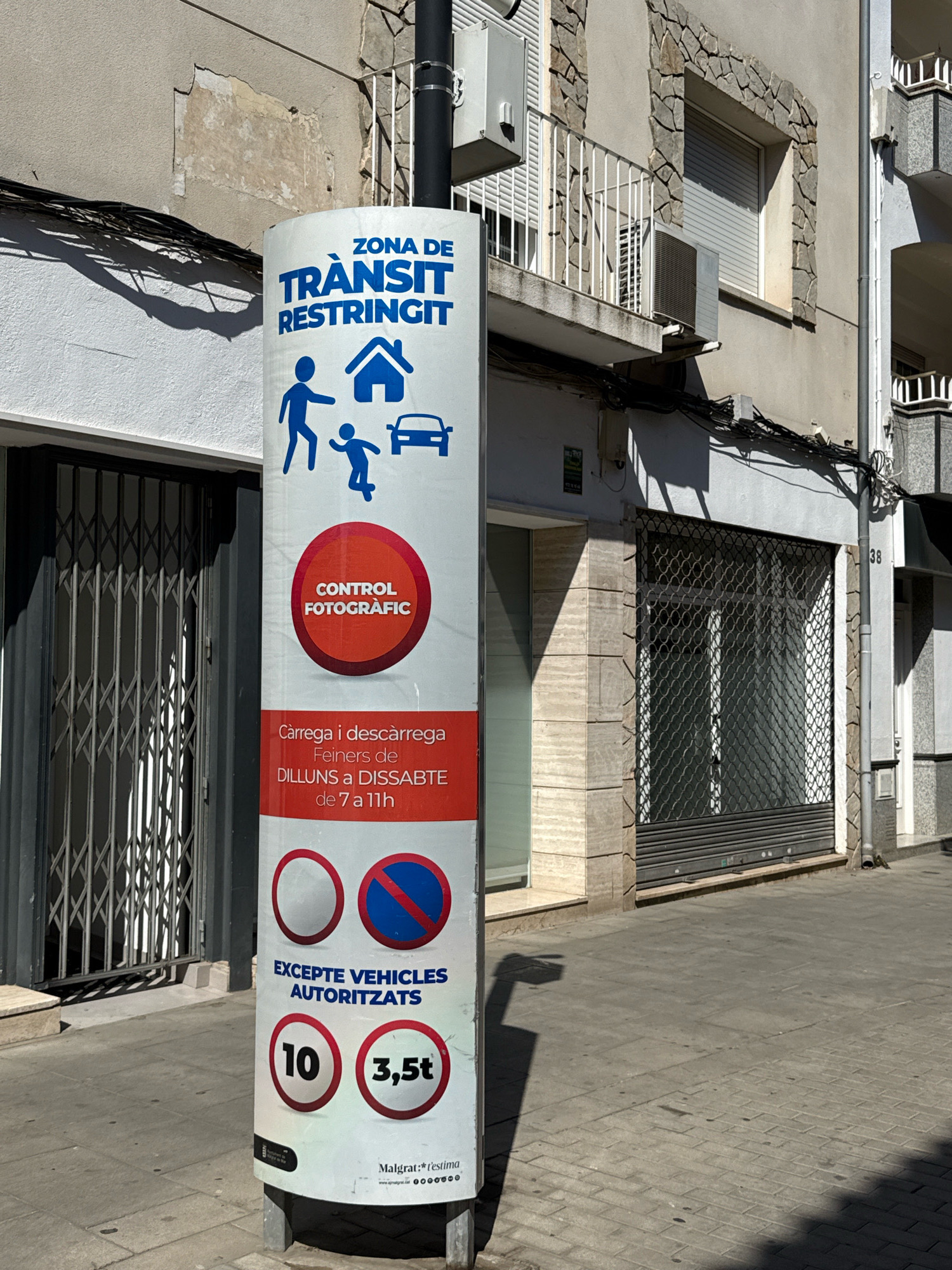 The image depicts a street scene in Malgrat de Mar, Spain. A cylindrical signpost stands prominently in the foreground, displaying information about restricted traffic zones. The signpost is white with blue and red text and icons. At the top, it reads "ZONA DE TRÀNSIT RESTRINGIT" (Restricted Traffic Zone), followed by icons of a person walking with a child and a house, and a car. Below this, a red circular section states "CONTROL FOTOGRÀFIC" (Photographic Control). Further down, it details "Càrrega i descàrrega Feiners de DIJOUS a DISSABTE de 7 a 11h" (Loading and unloading workers from Thursday to Saturday from 7 am to 11 am). Two traffic signs are displayed: a white circle and a blue circle with a red diagonal line, indicating prohibitions. Below these, it says "EXCEPTE VEHICLES AUTORITZATS" (Except authorized vehicles). Two weight limit signs are shown: one with "10" inside a circle and another with "3,5t". The bottom of the sign displays "Malgrat:*l'estima" and a website address.
The signpost is positioned on a paved sidewalk, with cobblestones visible in the lower right section. To the left of the signpost, a dark metal accordion gate is visible, suggesting an entrance to a building. The background shows a building with a stone facade, windows with blinds, and a balcony with a white railing. An air conditioning unit is mounted on the exterior wall. Electrical wires are strung along the building's facade. Sunlight casts shadows across the scene, indicating a bright, sunny day. The overall environment appears to be an urban street, possibly a commercial or residential area.