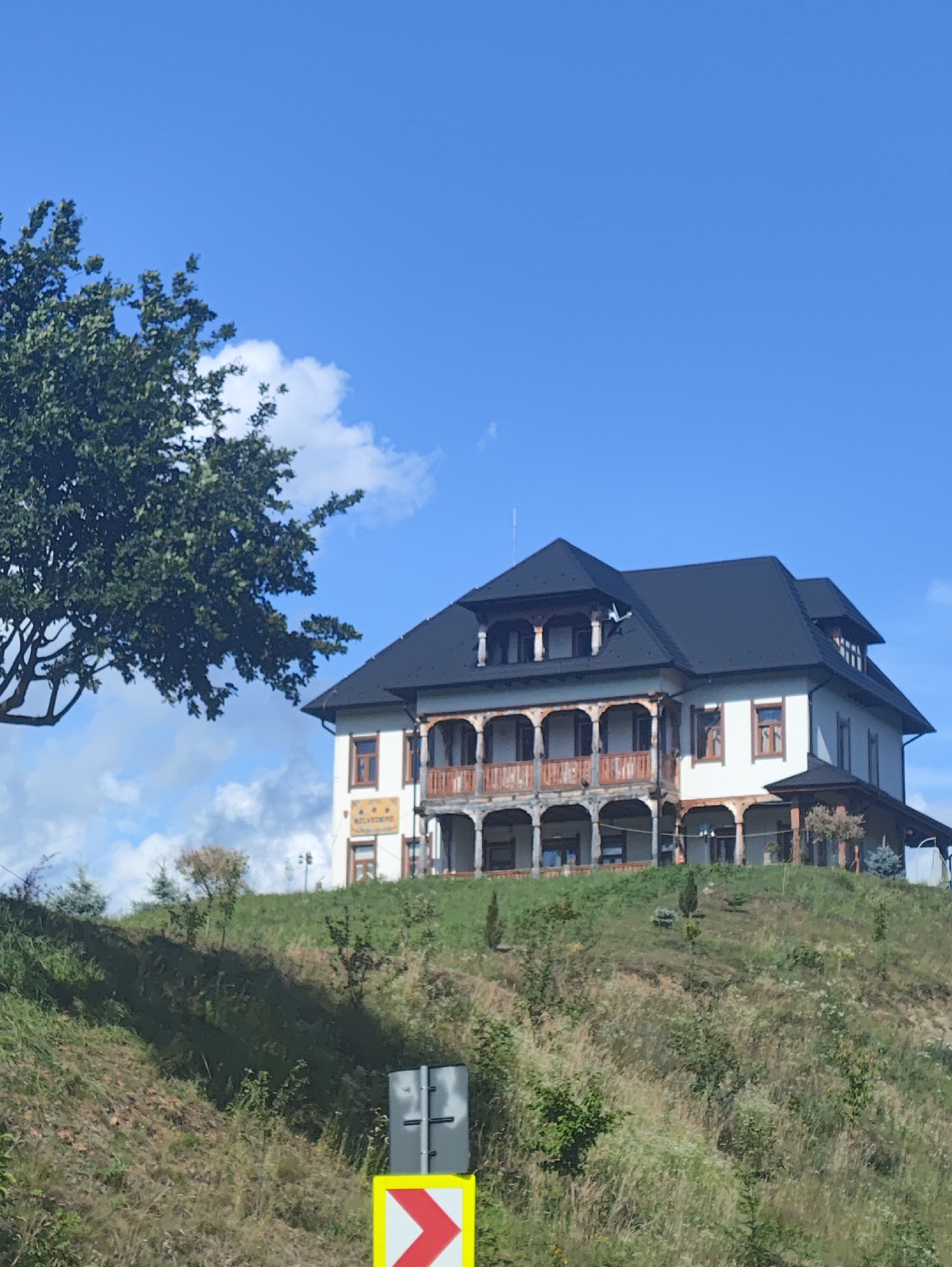 The image shows a white building with a dark roof, featuring multiple balconies with wooden railings, situated on a grassy hillside under a clear blue sky. The building has several windows and appears to be a hotel or guesthouse, with a sign that reads "BELVEDERE". A large tree with green leaves is visible to the left of the building. In the foreground, a road sign with a yellow background and a red chevron indicates a curve to the right. The environment is natural and scenic, with rolling hills covered in grass and vegetation. No people are visible in the image. The scene is likely located in Petrova, Romania, given the context.