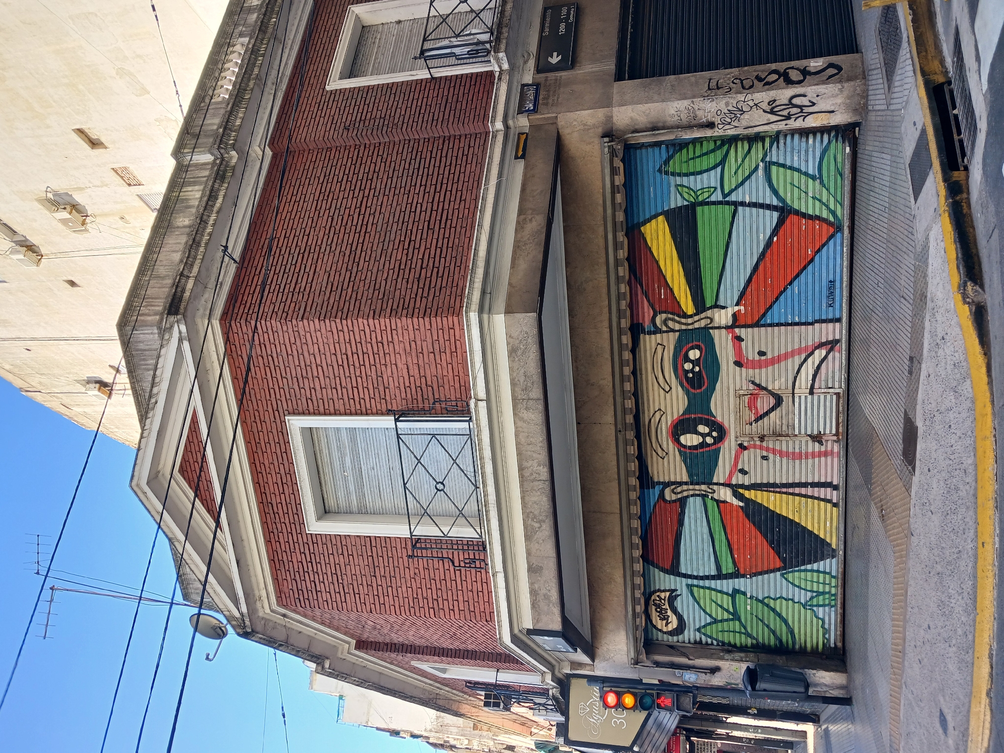 A brick building in Buenos Aires, Argentina, has a closed metal garage door with colorful street art of a face with leaves, and a street sign above it indicating Sarmiento 1200-1100, Corrientes 1. A traffic light shows red. The building has closed window shutters and security bars. The sky is blue.