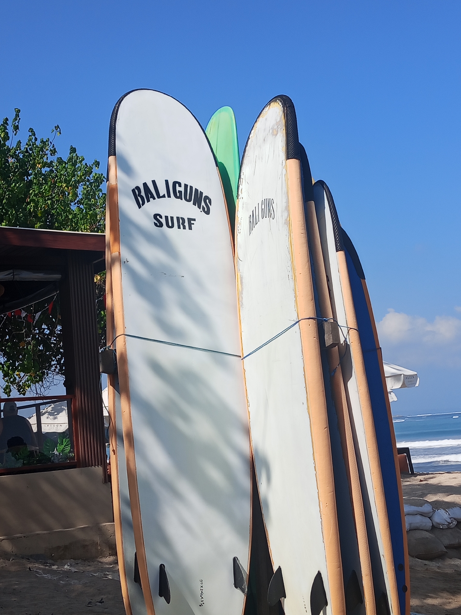 A row of surfboards stands vertically on a sandy beach in Kuta, Indonesia, under a clear blue sky. The surfboards are leaning against each other, with the branding "BALIGUNS SURF" visible on the foremost white board, along with smaller text on subsequent boards. A green surfboard is positioned behind the white one. To the left, a section of a wooden structure with a roof casts shadows on the surfboards. In the background, the ocean with gentle waves meets the horizon, and a white beach umbrella with a sign that appears to read "LAGOO..." is visible. Several sandbags are stacked near the water's edge. The scene depicts a typical day at a surf spot in Kuta.