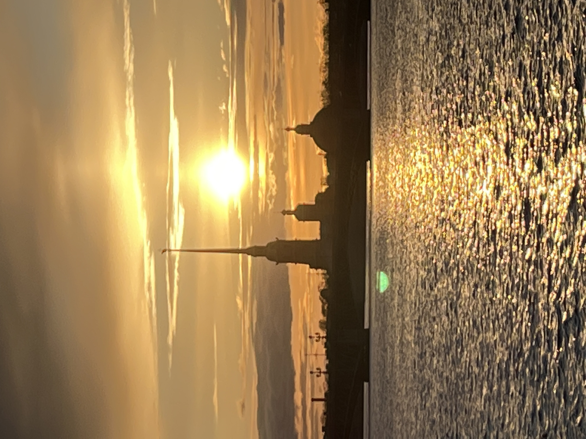 The image shows a golden sunset over the Neva River in Saint Petersburg, Russia. The silhouette of the Peter and Paul Fortress, including its distinctive golden spire, and other buildings are visible against the setting sun. The water reflects the golden light, creating a shimmering effect.