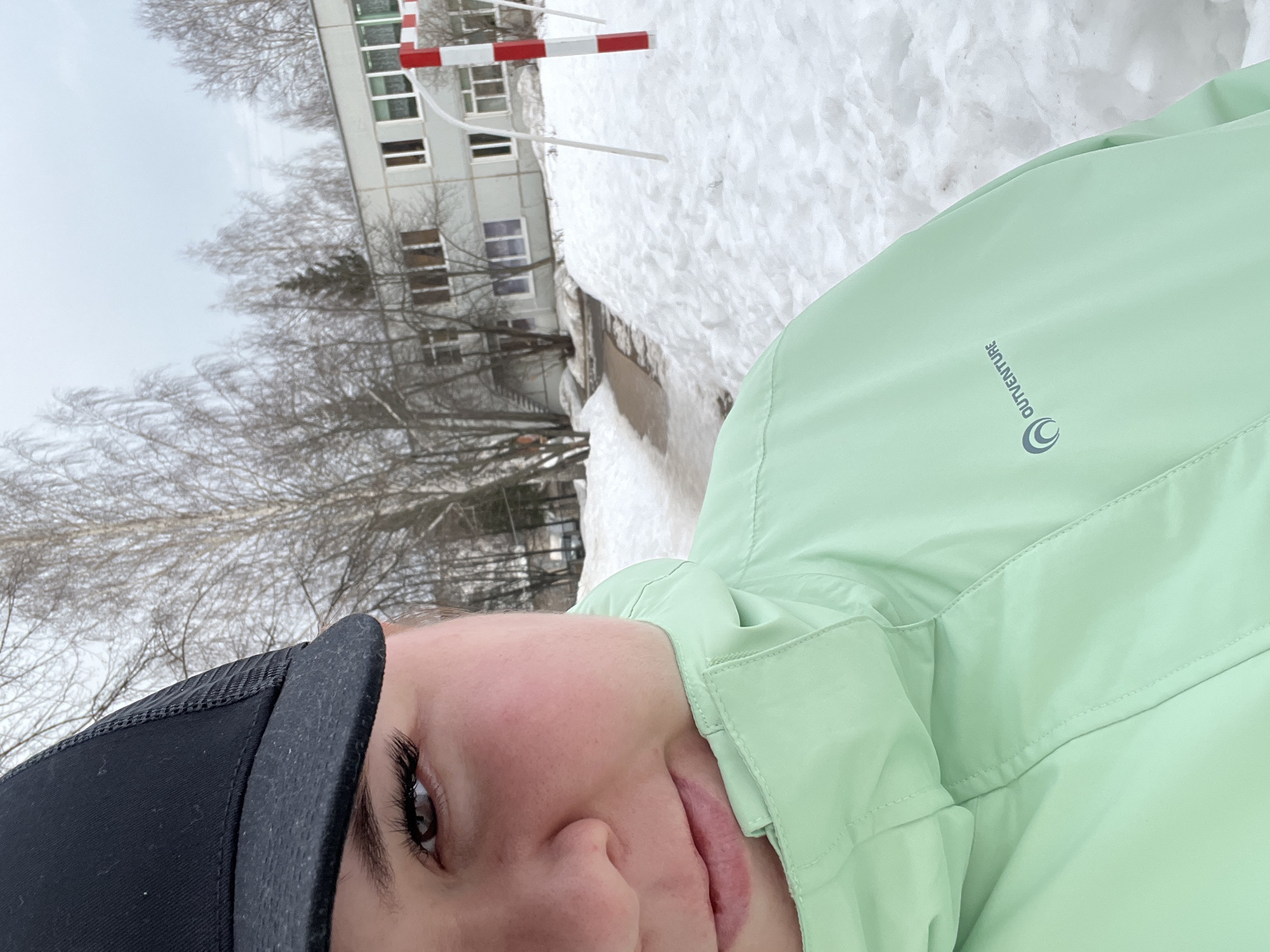 The image displays a close-up, chest-up shot of an individual, likely female, in an outdoor, snow-covered setting in Chaikovsky, Russia. The person's right eye, part of the nose, and mouth are visible on the left side of the frame. The eye is dark brown with prominent eyelashes. A black baseball cap with a mesh back covers the upper portion of the head. The individual is wearing a light mint green winter jacket with a high collar and a zipper closure. The right side of the jacket features a grey "OUTVENTURE" logo with a stylized crescent graphic.
The background shows an overcast winter or early spring landscape. Extensive snow covers the ground, with a partially cleared or melted pathway curving through it on the left. In the mid-ground, a multi-story, light-grey building with numerous windows is visible, partially obscured by bare deciduous trees and one dark evergreen tree. On the right, a red and white striped pole, appearing to be part of a small goal or play structure, rises from the snow, accompanied by two thin white support poles. The sky is uniformly light grey, indicating an overcast condition.