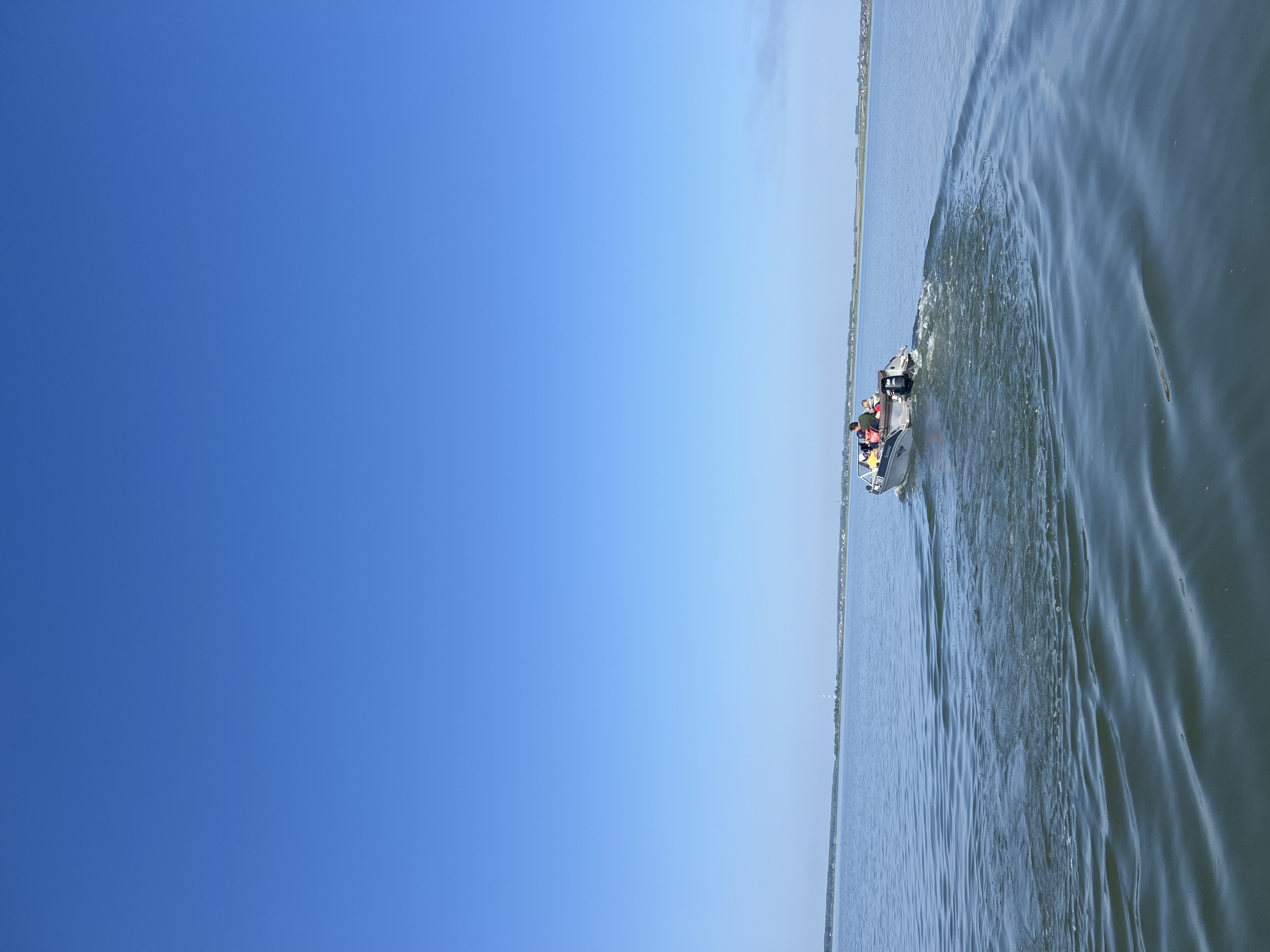An eye-level, outdoor, vertical shot shows a clear blue sky over a body of water in Novopichugovo, Russia. The water is calm with small ripples, and a wake emanates from a small, silver motorboat with three people on board. The boat is moving away from the viewer towards the right side of the frame. The background shows a distant shoreline with some buildings and vegetation. The lighting suggests it is daytime with clear weather.