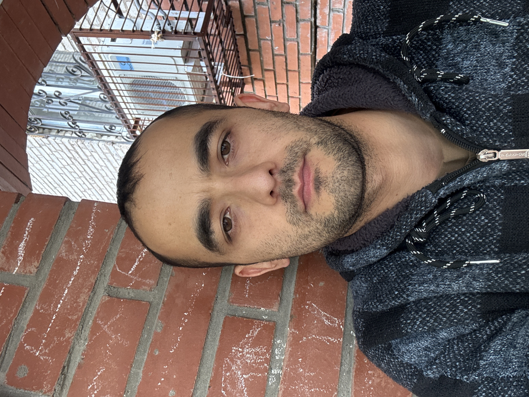 A man with short dark hair and a beard, wearing a dark hooded sweater, is captured in a close-up, head-and-shoulders selfie. He is positioned to the left of the frame, leaning against a red brick wall, with his body angled towards the camera. The background reveals a building with white brickwork, a window with decorative metal bars, and an air conditioning unit mounted on a metal grate. The lighting suggests an outdoor setting during the daytime. The context of Ufa, Russia, is not visually ascertainable from the provided image.