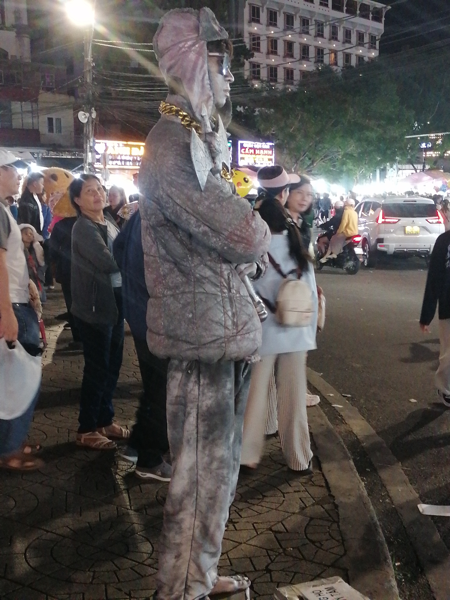 The image depicts a street scene at night in Da Lat, Vietnam, likely during a public event or festival. In the foreground, a person in a full-body costume, appearing to be a street performer, stands facing right. The costume is predominantly silver-gray, with a large gold chain around the neck. The performer wears sunglasses and a hood that covers the head. The costume's texture suggests it's made of padded fabric.
To the left of the performer, several people are gathered on a patterned tiled sidewalk. One woman in a dark jacket and jeans stands with her arms crossed, looking towards the performer. Another individual in a white hooded jacket is partially visible behind her. Further left, a person in a white t-shirt and blue jeans holds a white plastic bag.
In the mid-ground, more people are visible, including a woman in a light blue jacket and striped pants, carrying a small beige backpack. Behind her, a scooter with two riders, one wearing a yellow jacket, moves along the street. A white SUV is also present on the road. The background shows buildings, trees, and streetlights, indicating an urban environment. The overall lighting suggests it is nighttime, possibly with artificial illumination from street lamps and buildings. The presence of many people and the festive atmosphere hint at a lively public space in Da Lat.