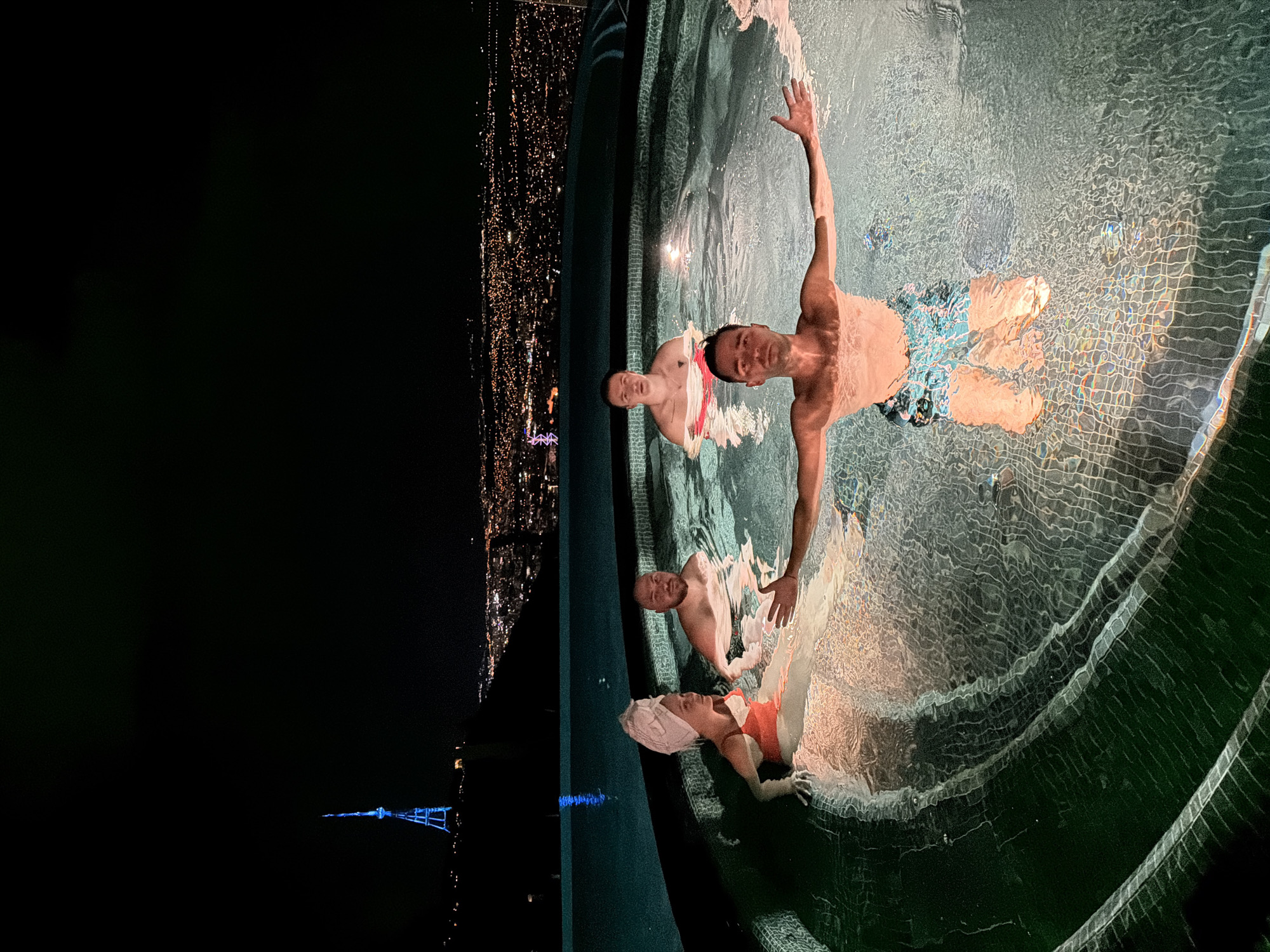 Three individuals are visible in a swimming pool at night, appearing to be relaxing or swimming. The pool is on a high vantage point, with the cityscape of Tbilisi, Georgia, visible in the background. The city lights create a bright contrast to the dark night sky. One of the individuals is standing in the water with an arm raised. The individual in the lower part of the frame is wearing an orange bathing suit and a white head towel. A third individual is partially visible in the pool between the other two. The pool's edge is tiled.
