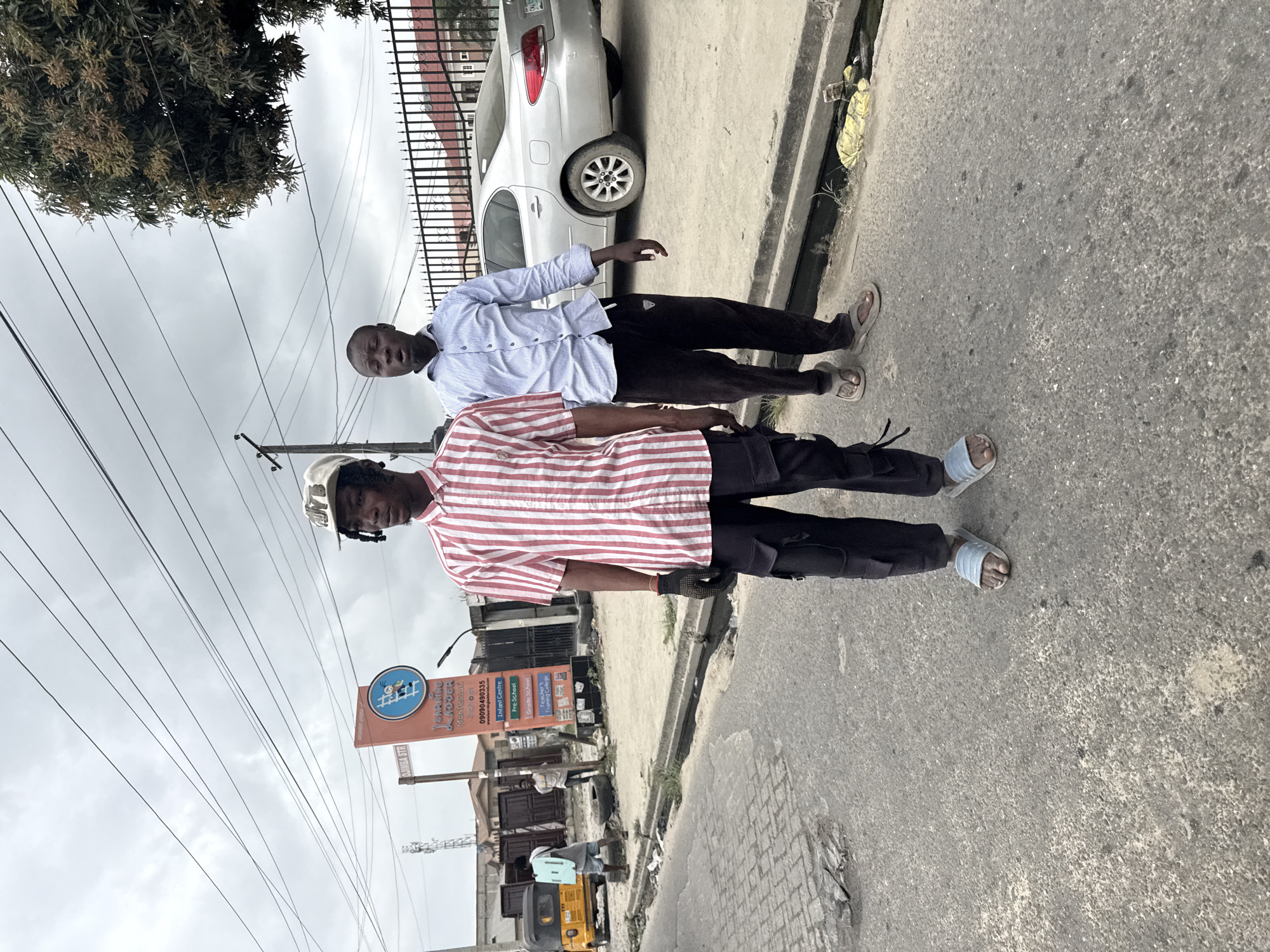 In Ado, Nigeria, two young men stand on a paved road under an overcast sky. The man in the foreground wears a red and white striped short-sleeved shirt, black cargo pants, and light blue slides. He has black dreadlocks and wears a light-colored baseball cap. He is looking to his left. To his right and slightly behind him, another young man stands wearing a light blue striped long-sleeved shirt and dark pants with brown slides. His mouth is open, as if speaking.
Behind them, a silver sedan is parked. A black metal fence with decorative posts encloses a property with a red-tiled roof visible behind the fence. A signpost with the words "LEARNING LADDER Montessori School" and a phone number is also visible. To the left of the sign, a yellow auto-rickshaw is parked. Two other individuals are visible further down the road near the auto-rickshaw, one carrying a light blue object on his back. Utility poles and overhead wires crisscross the sky. The road has patches of disrepair and a narrow drainage ditch runs along the side, containing some debris.