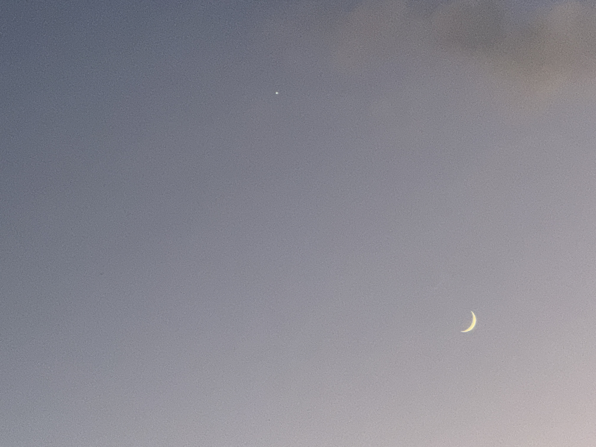 The image is a photograph of the sky at dusk over Adeje, Spain. A small, bright point of light, likely a planet or star, is visible in the upper portion of the frame. In the lower right quadrant, a thin crescent moon is visible. The sky is a gradient of muted blues and purples, with a faint warm glow on the right horizon, suggesting the last vestiges of sunlight. There are no people, structures, or other discernible objects in the image, which is dominated by the celestial bodies and the twilight atmosphere. The image has a grainy texture, indicative of low-light photography or processing.