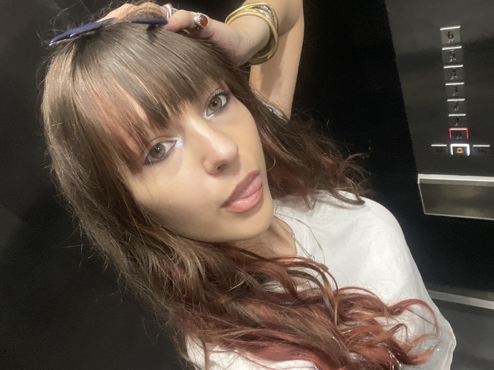 A young woman with brown hair, styled with bangs and some reddish-pink highlights, is looking into the camera with her tongue slightly sticking out. She is wearing a white t-shirt and a few necklaces, and several rings and bracelets on her right hand. Her makeup includes white eyeshadow. Her right hand is resting on the side of her head, and her left arm is bent. In the background, a dark, indistinct wall is visible, with a panel featuring buttons and illuminated numbers in what appears to be an elevator. The scene is indoors and appears to be a dimly lit environment.