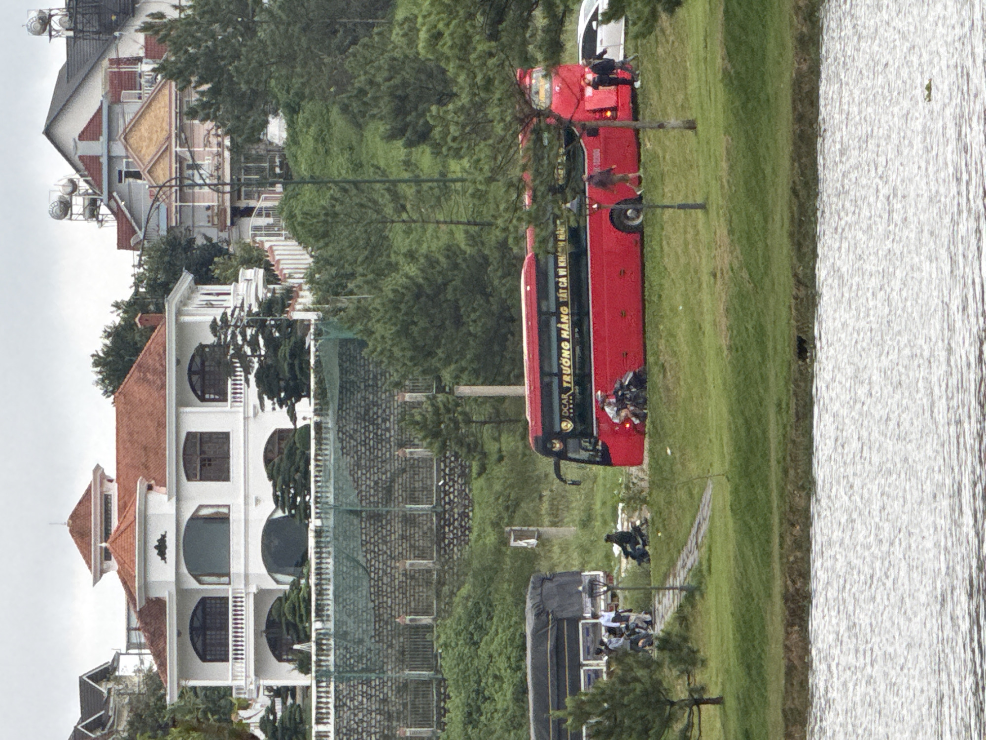 The image depicts a panoramic view of Xuân Hương Lake in Đà Lạt, Vietnam, under a cloudy sky. The foreground features the rippling surface of the lake. In the middle ground, a paved pathway runs alongside a grassy bank, leading to a road where several vehicles are present. A large red bus with Vietnamese text on its side is parked, with several people standing near it. A black truck is also parked nearby. Several individuals are seen walking or standing around the vehicles. Further in the background, a multi-story white house with red-tiled roofs is visible on a hill, surrounded by lush green trees. Other houses with similar architectural styles are also present on the hillside. The overall scene captures a moment of activity by the lake, with transportation and residential elements in the backdrop.