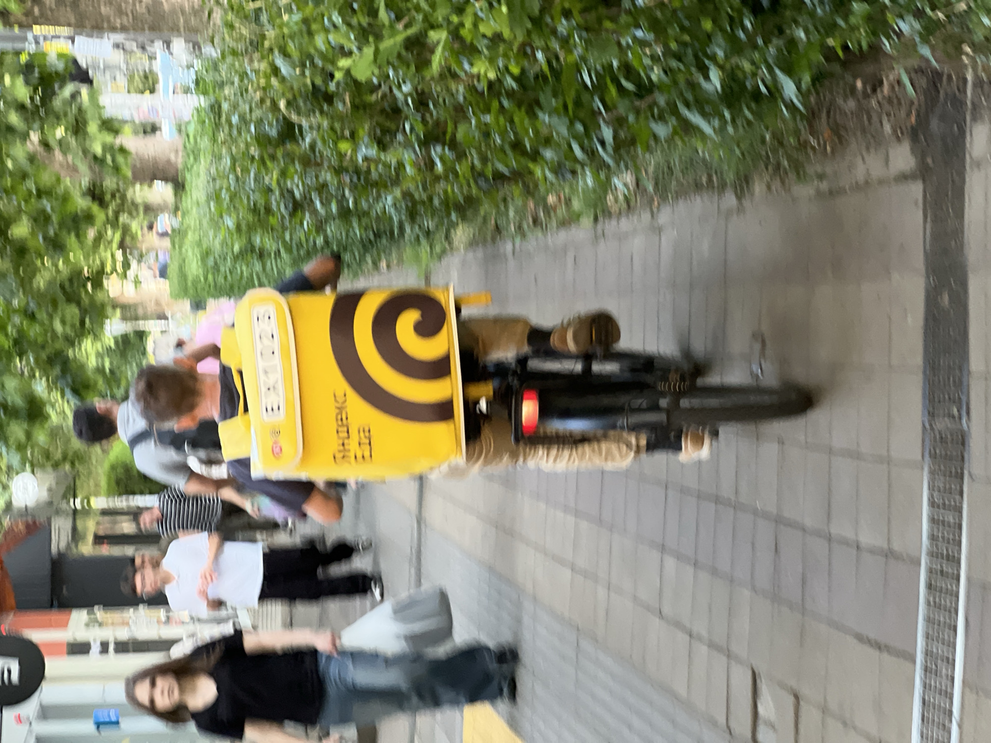 The image depicts a delivery person on a bicycle, riding on a pedestrian sidewalk in the city of Krasnodar, Russia. The delivery person is wearing light-colored pants and has a yellow insulated backpack on their back, with the branding "Яндекс Еда" (Yandex Food) visible. The backpack has a distinctive brown spiral design. In the background, several other people are walking on the sidewalk, and there are trees and buildings lining the street. The sidewalk is paved with gray tiles. The lighting suggests it is daytime, possibly late afternoon. The scene appears to be a typical urban environment with pedestrian and vehicular traffic.