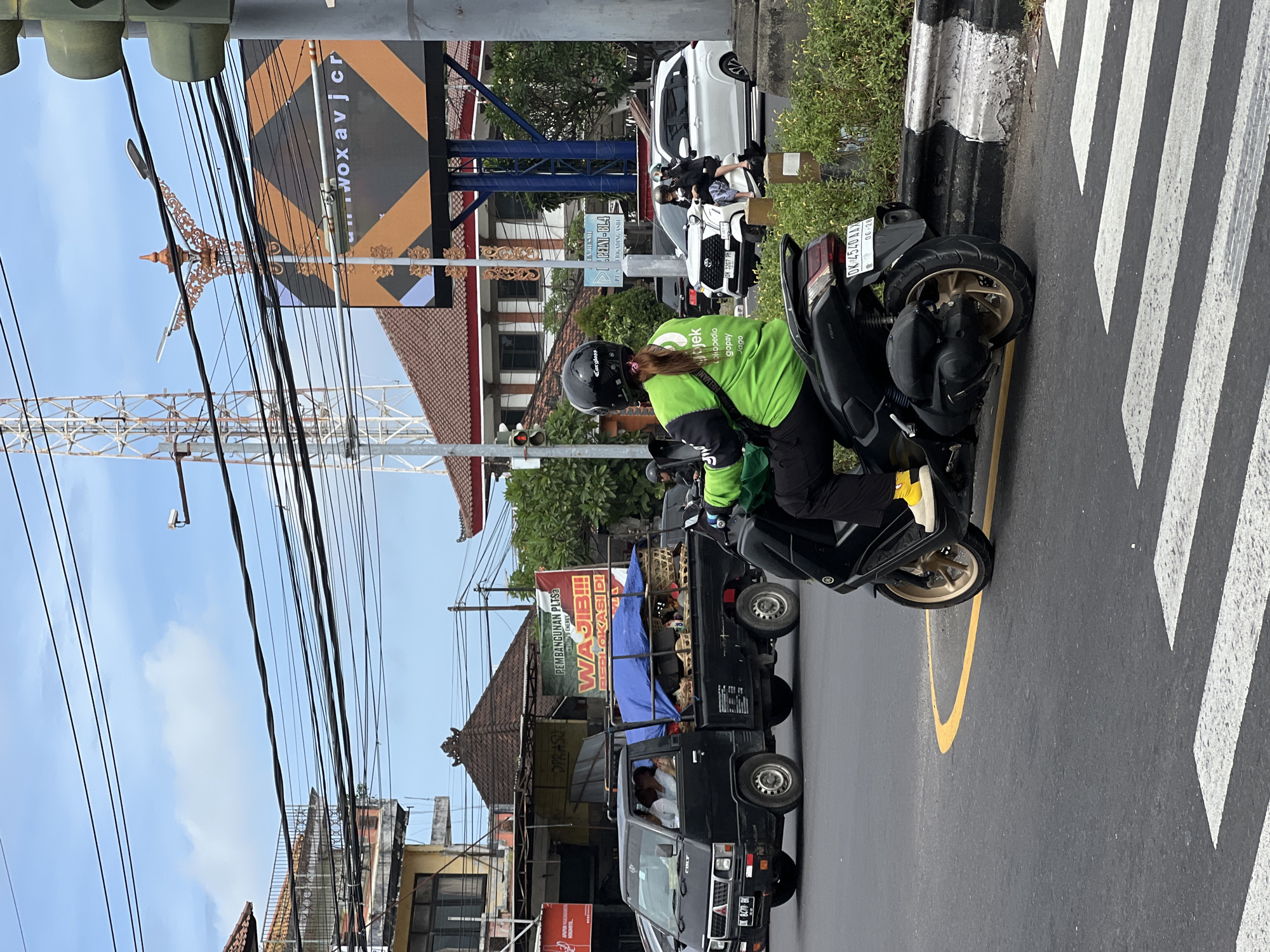 The image depicts a street scene in Denpasar, Indonesia. A woman wearing a green Gojek uniform and a black helmet is seated on a black motorcycle facing away from the camera. The motorcycle is stopped on a road marked with white and yellow lines. A black pick-up truck with a blue tarp and goods on the back is in the lane beside the motorcycle. Overhead, there are numerous power lines, a traffic signal, a tower structure, and a billboard that says "woxavjcr". Buildings with Balinese-style roofs are in the background. Street signage indicates "PEMBANGUNAN PLTSa" and "WAJIB!!! BERILOKASI DI". A white car is partially visible further down the road.