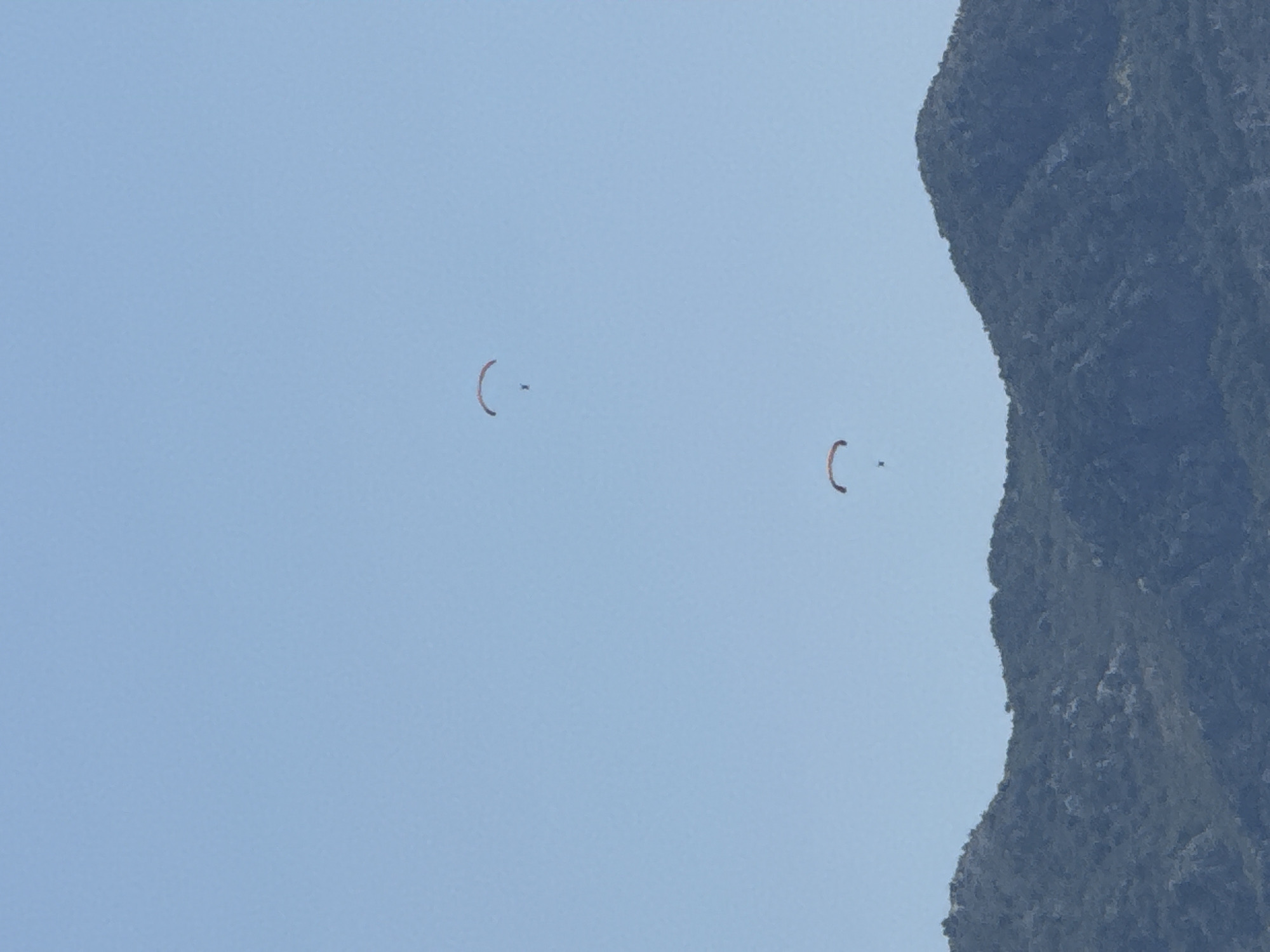 Two paragliders are depicted against a clear blue sky, soaring over a lush, green, mountainous terrain. The paragliders, one above the other, are positioned in the upper half of the image. The lower half of the image is dominated by a dark, textured mountain range, suggesting a natural, possibly remote, environment. While the image does not provide specific landmarks of Budva, Montenegro, the presence of mountains and clear skies is consistent with the geographical features found in the region. The paragliders are likely engaging in recreational activity above the coastal mountains.