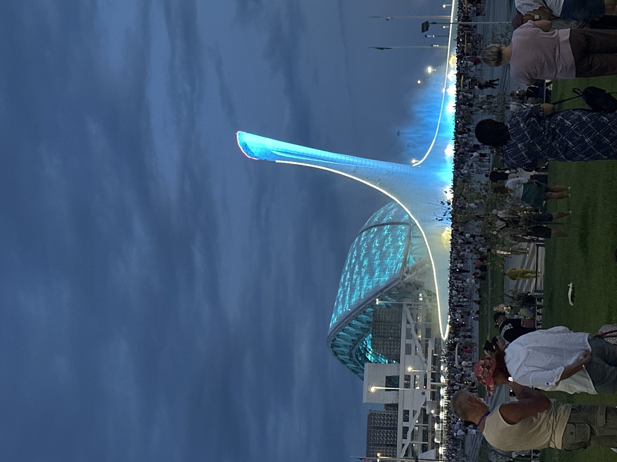 The image depicts the Fisht Olympic Stadium in Sirius, Russia, at dusk. The stadium's distinctive roof, illuminated with vibrant turquoise lights, is visible on the left. To the right, a tall, flame-like structure, also illuminated with blue and white lights, rises into the dark blue, cloudy sky. In the foreground, a crowd of people is gathered on a grassy area. Several individuals are the main focus in the foreground, including a man and a woman conversing, and two women facing away from the camera, observing the spectacle. The overall atmosphere suggests an outdoor event or attraction taking place in the evening.