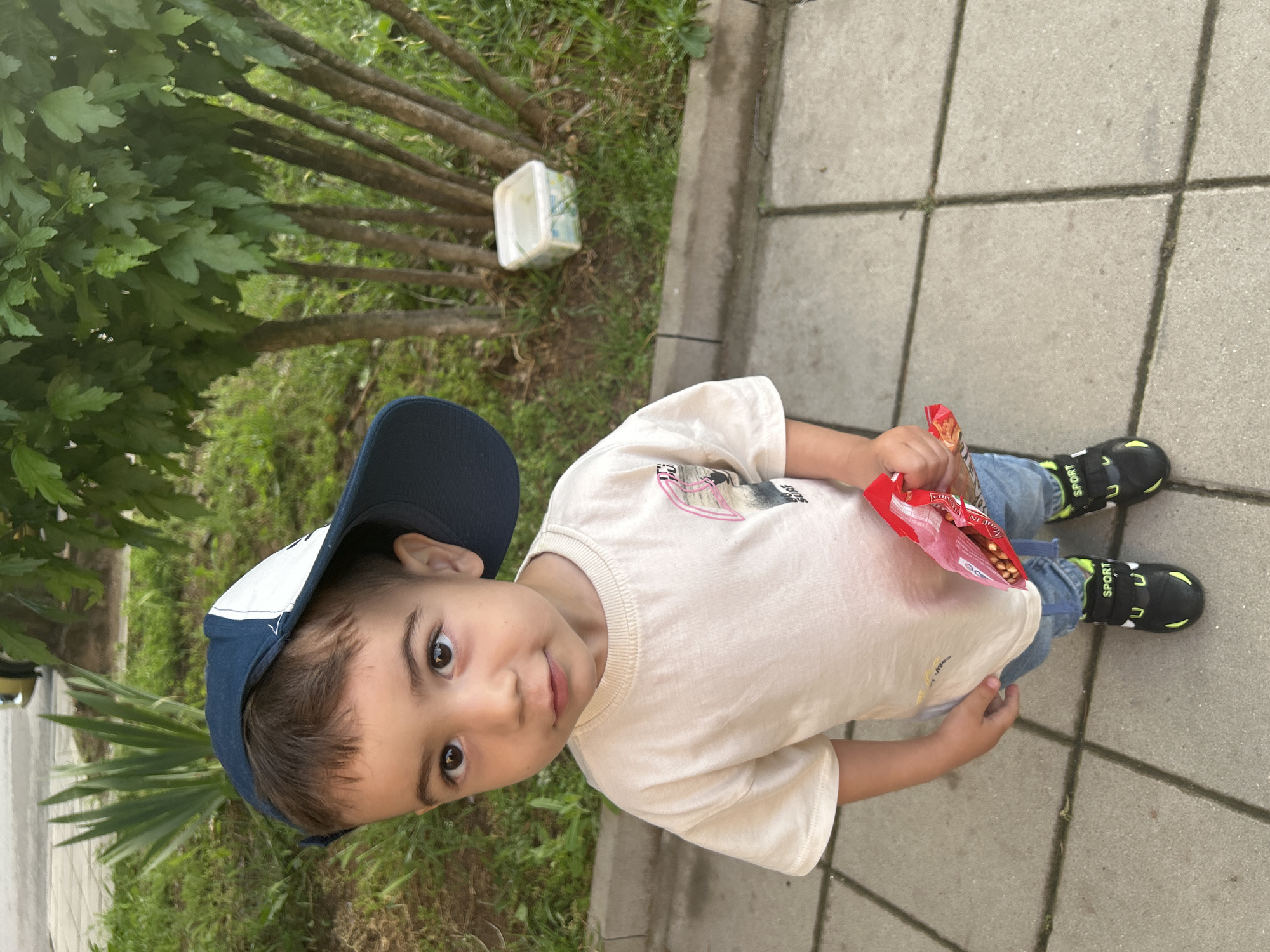 A young male child, approximately two to three years old, stands on a concrete tiled sidewalk in Kardzhali, Bulgaria. He has light brown hair, dark brown eyes, and fair skin, looking directly at the viewer with a neutral expression. He is wearing a beige short-sleeved t-shirt with a partially visible graphic design on the left chest, blue jeans, and black athletic shoes with lime green accents and "SPORT" written on the velcro straps. A dark blue baseball cap with a white front panel is worn backward and askew on his head. The child holds a translucent red plastic bag containing small, light brown, spherical snack items in his right hand. To the child's left and behind him, a grassy area with various green plants and slender-trunked trees is present, separated from the sidewalk by a concrete curb. A white plastic container, partially filled with liquid, rests on the ground amidst the foliage behind the child. A small section of a yellow vehicle is minimally visible in the upper left background.