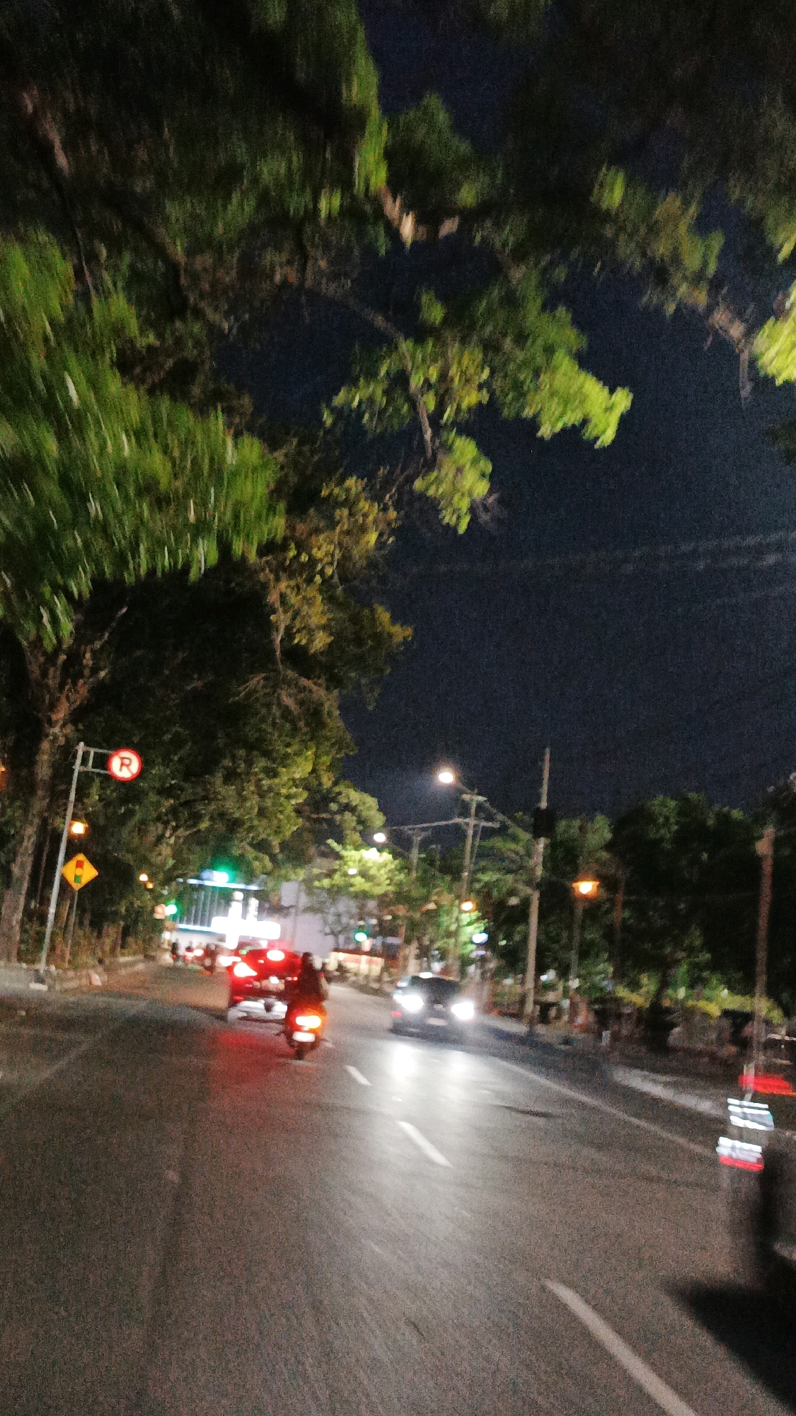 The image shows a blurred, nighttime scene on a city street in Surakarta, Indonesia. A motorcycle with its taillight illuminated is traveling on the asphalt road. To the left, there is a traffic sign with a red circle and a white "R" inside, indicating no parking. Adjacent to it is a yellow diamond-shaped sign with a black arrow pointing upwards. Further down the road, streetlights and the headlights of a car are visible. Trees with green foliage line the street. The sky is dark, suggesting it is nighttime. The overall impression is that of a typical urban environment at night in Surakarta.
