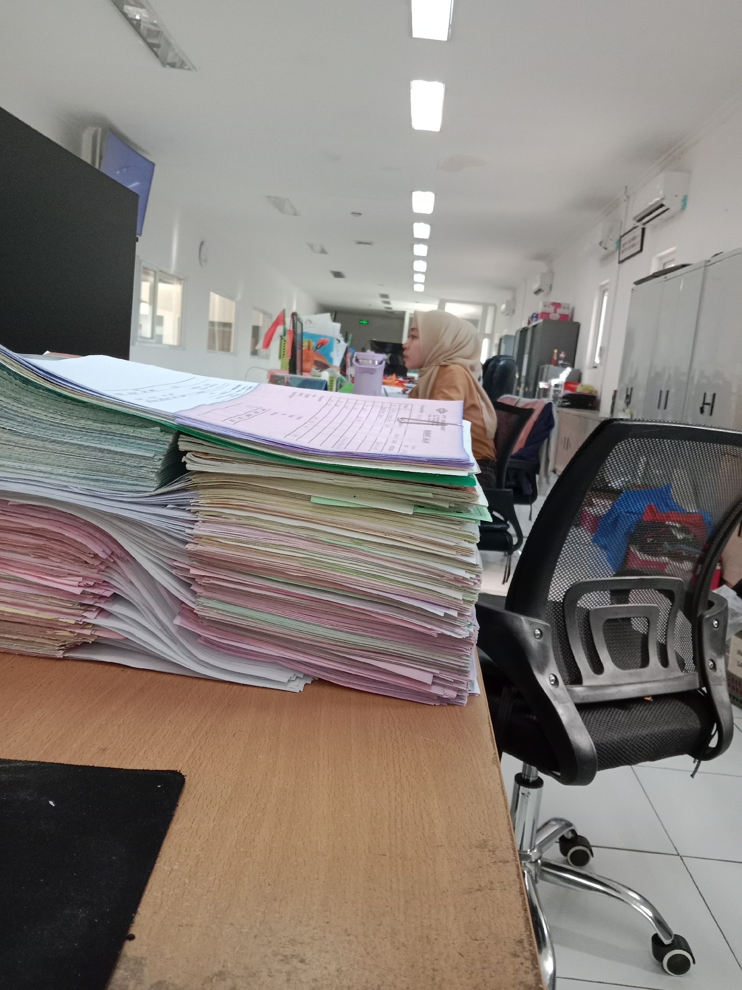 A woman wearing a hijab is seated at her desk in an office, with a large stack of documents in the foreground. The office environment appears to be modern, with white walls, ceiling lights, and air conditioning units visible. Filing cabinets and office chairs are also present. The overall scene suggests a typical workday in an Indonesian office, possibly in Semarang, Indonesia, given the context of the query.