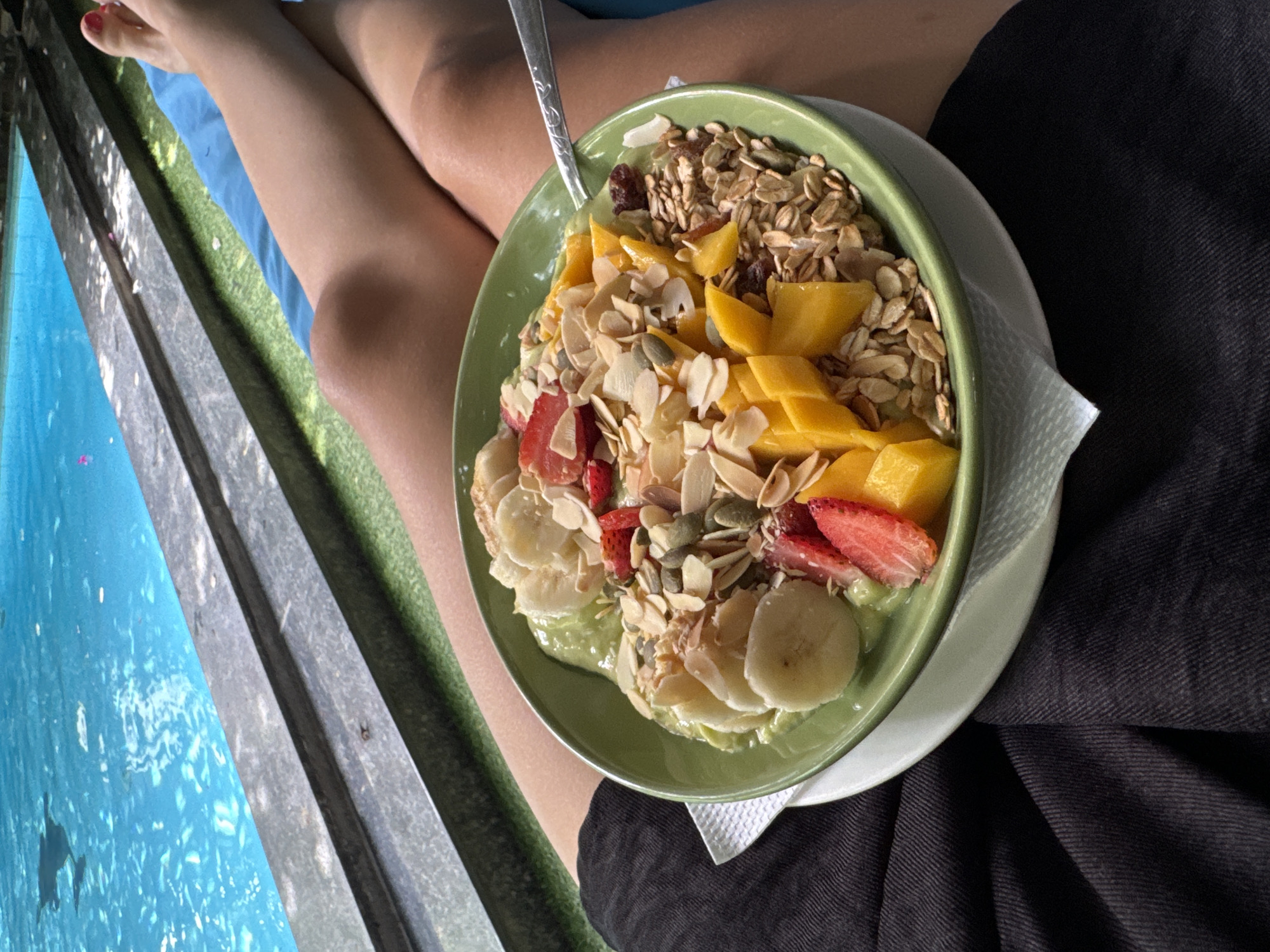 The image shows a person holding a green bowl filled with a smoothie and various toppings. The smoothie is green and topped with sliced bananas, sliced strawberries, mango pieces, granola, and sliced almonds. A spoon rests in the bowl. The person's legs are visible, and the bowl rests on their lap. To the left of the person, there is a swimming pool with clear blue water and a gray, metal fence. The person is wearing black pants.
Given the prompt and the image, it is impossible to verify whether or not the location of the image is Mũi Né, Vietnam. No distinguishing landmarks or signs indicate the location of the image.