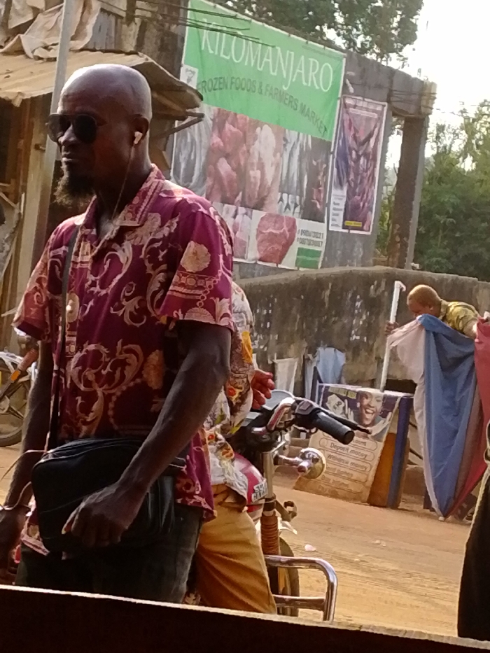This image depicts an outdoor scene in Enugwu-Ukwu, Nigeria, likely a market or commercial street area.
In the left foreground, an adult male with dark skin and a bald head is visible from the chest up, facing right. He wears dark sunglasses and white wired earbuds. A short, dark beard is visible on his chin. He is dressed in a short-sleeved, collared shirt with a maroon/burgundy base featuring intricate gold and beige ornate patterns. A black shoulder bag is slung across his body, with his left hand resting on it. He appears to be either standing or walking alongside a motorcycle, a portion of which is visible to his right, including chrome handlebars.
Behind the prominent male subject, another individual is partially visible, wearing lighter-colored patterned clothing, seated on what appears to be the motorbike.
The background features a concrete wall or building facade adorned with multiple advertising banners. A large green banner prominently displays "KILOMANJARO FROZEN FOODS & FARMERS MARKET," with images of raw meat and possibly fish. To its right, a smaller purple banner also depicts merchandise. Further right, a person with light-colored hair, wearing a patterned shirt, is partially obscured by blue and white fabric, appearing to interact with merchandise or a stall, holding a white pole. Below this, another sign is visible with a smiling face and text including "Deposit money" and "Withdraw money," suggesting financial services advertising. The ground is unpaved and dusty. Sparse vegetation is visible in the distant background on the right.