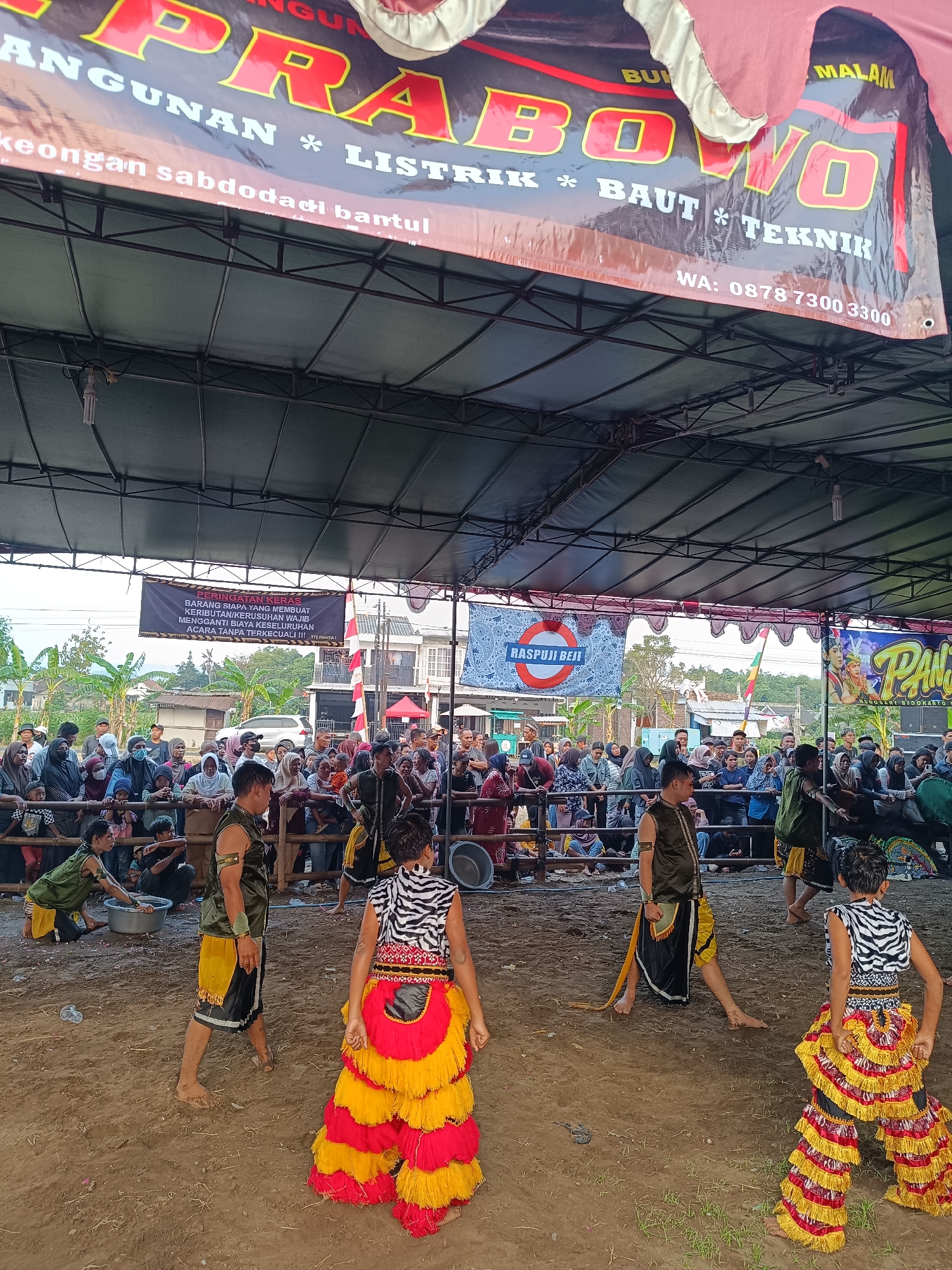 This is a medium shot, eye-level outdoor photograph of a performance event taking place in the city of Jetis, Indonesia. The foreground is dominated by a dirt ground with scattered grass. In the center of the frame, four individuals in traditional performance costumes are visible. One performer in the foreground right wears a black and white zebra-striped top and heavily fringed red and yellow pants. To their left, another performer in a black and gold outfit walks away from the camera. To the left of them, a child in a green and yellow outfit is kneeling and interacting with a metal basin. Further to the left, another performer in a similar red and yellow fringed costume is partially visible.
In the mid-ground, a crowd of spectators is gathered behind a wooden barrier, many wearing headscarves and casual clothing. Several banners are displayed above the performance area and the crowd. One banner in the upper portion of the image features large red and yellow text. Another black banner displays text in white and red, including a warning notice and a phone number. To the right of the crowd, a colorful banner with images of two figures is visible.
The background shows a mix of buildings, trees, and utility poles, indicating a semi-urban or rural setting. The overall lighting suggests an overcast or late afternoon sky. The image captures a moment during a cultural performance, with performers and spectators present.