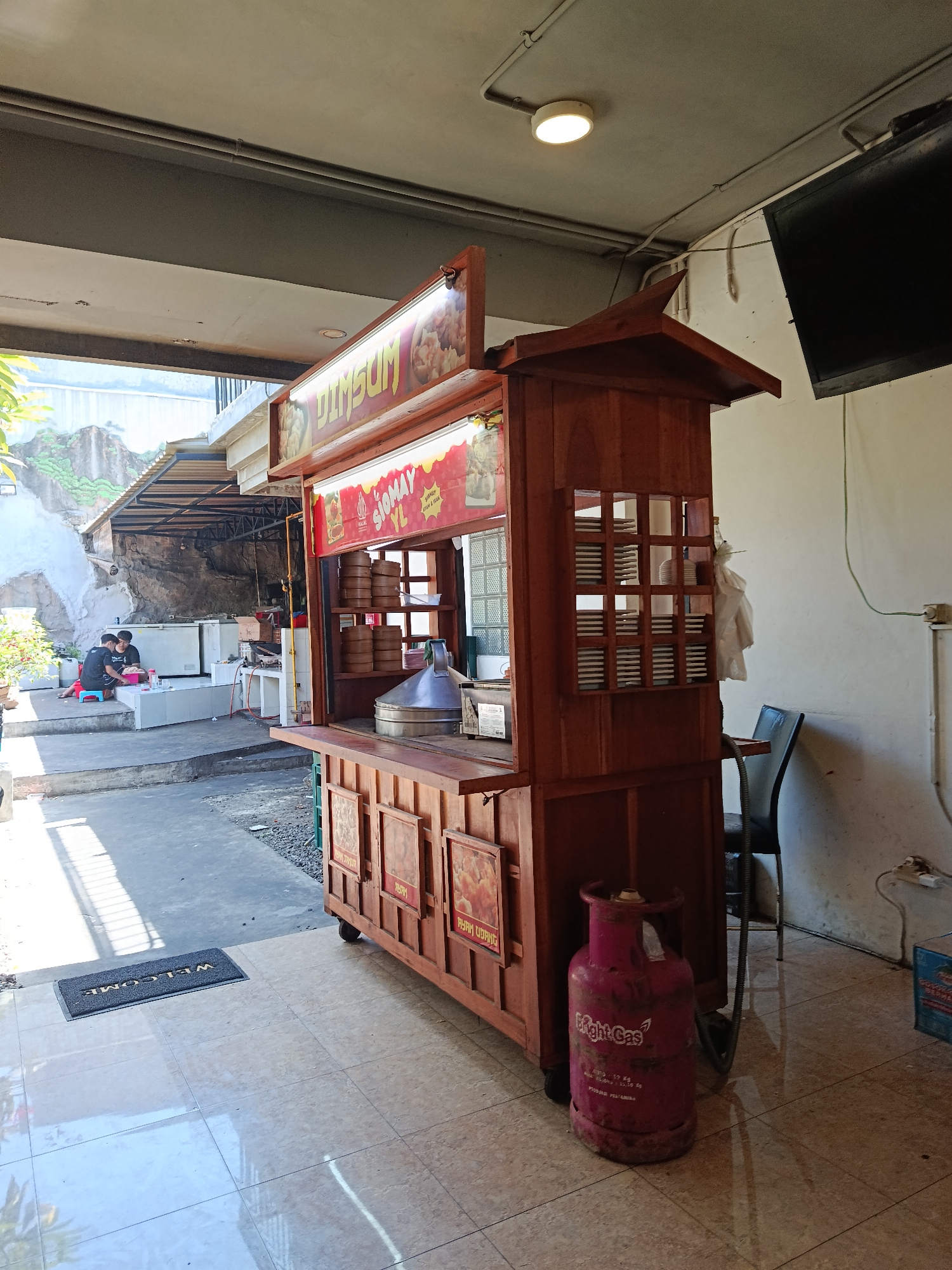 The image depicts a street food stall named "DIMSUM" in a bright, open-air setting, possibly in Surabaya, Indonesia. The stall is made of wood and features a prominent display of dim sum steamers stacked on shelves. Above the counter, a colorful sign advertises "DIMSUM" and "SIOMAY YL," with accompanying images of food. To the right of the stall, a pink gas cylinder is visible on the tiled floor. In the background, two individuals are seated near refrigerators and what appears to be a preparation area. The environment suggests a casual dining or food service establishment.
