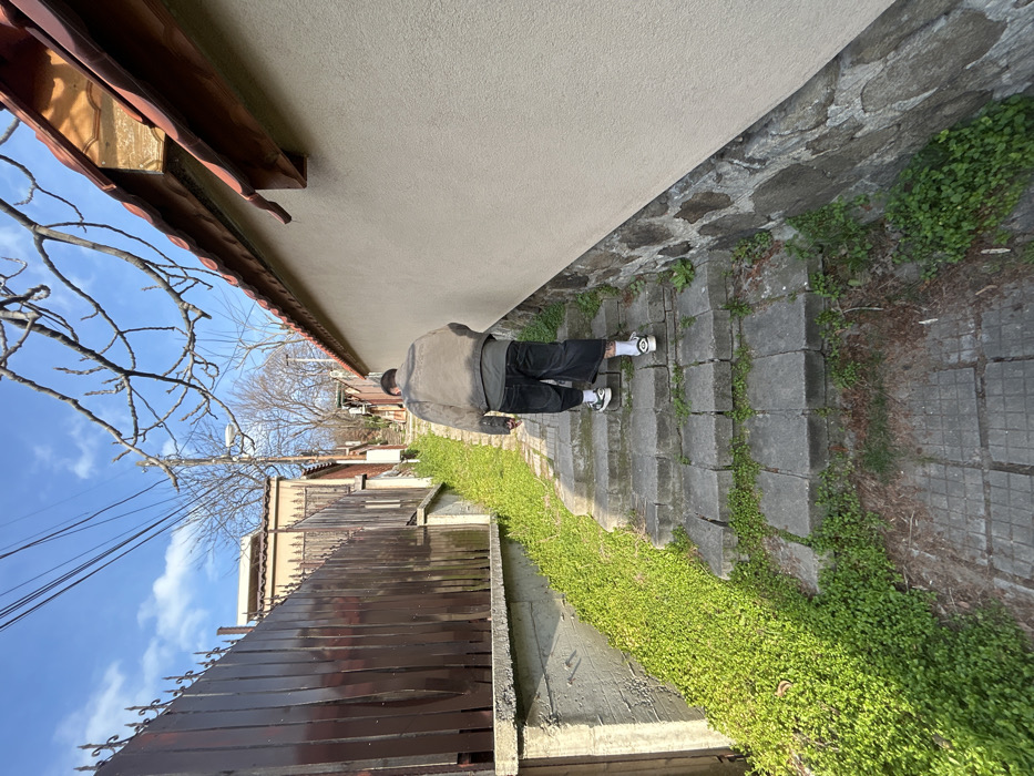 The image depicts an uphill pedestrian pathway in the Old Town area of Plovdiv, Bulgaria. A single individual is observed from behind, ascending a series of irregularly shaped stone steps. The subject is wearing a light-colored, possibly distressed, jacket over a dark t-shirt, dark mid-calf length shorts or cropped trousers, white socks, and dark sneakers with white soles. A tattoo is visible on the lower left leg.
To the left of the pathway, a concrete retaining wall supports a tall, dark brown wooden fence topped with decorative metal spikes. Beyond this fence, part of a building with a light-colored wall and a reddish-brown tiled roof is visible. Green ground cover densely occupies the sloped terrain between the retaining wall and the steps.
To the right of the pathway, a retaining wall constructed from irregular natural stones supports a building with a smooth, light-colored stucco wall. Above this, a roof with terracotta tiles and a wooden overhang is visible. Bare tree branches extend into the upper right portion of the frame.
In the upper central and left areas, utility poles with overhead wires are visible, along with a street light further up the path. The sky is clear blue with minimal clouds. The overall lighting suggests bright daylight.
