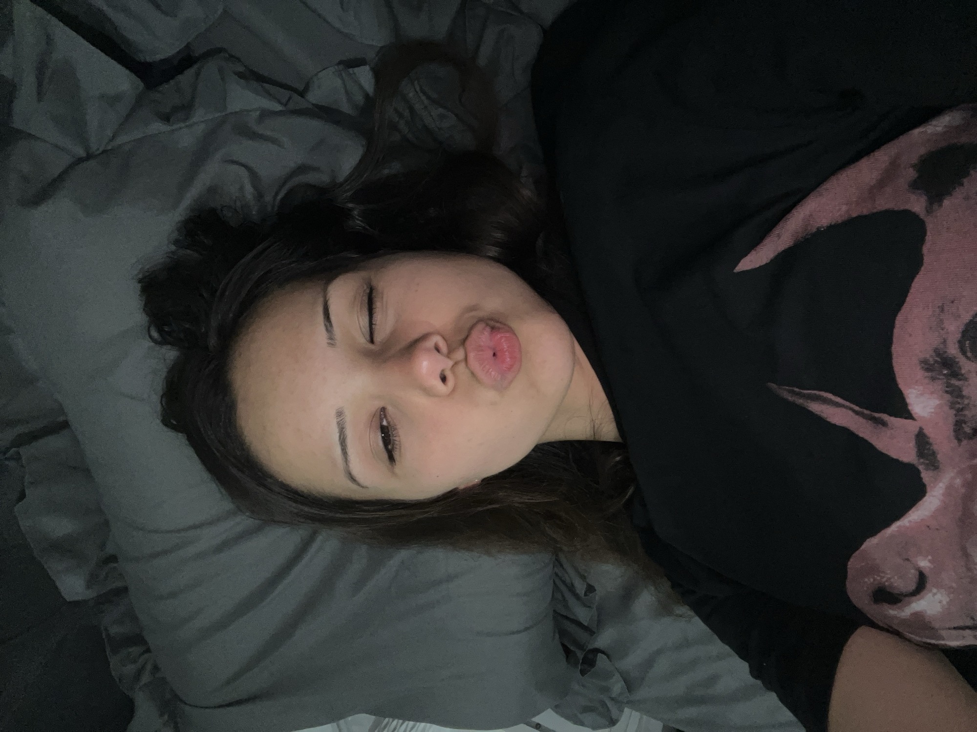 A young woman with dark hair lies on a dark gray duvet, her head resting on a pillow. She is wearing a black long-sleeved shirt with a large pink graphic design on the left side. Her eyes are closed, and her lips are pursed in a kissing gesture. Her right eyebrow is visible, and she has makeup on. The scene appears to be indoors, possibly a bedroom. The provided location of Zlatograd, Bulgaria, is noted but not visually represented in the image.