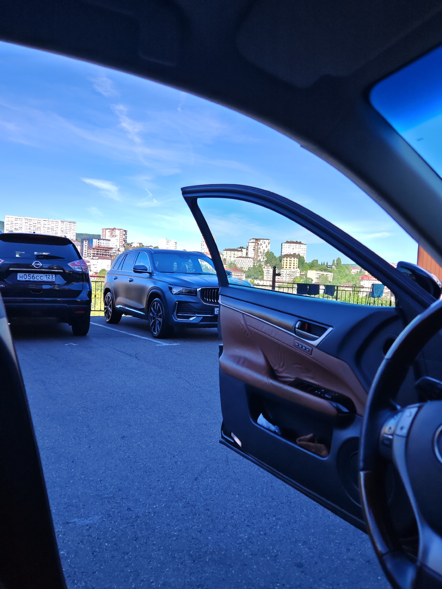 The image presents a daytime view from the interior of a car with its front door open. In the immediate foreground, the open door reveals a brown leather interior panel with various controls and a partial view of the steering wheel. The car is parked on an asphalt surface.
In the mid-ground, two SUVs are parked. On the left, the rear of a black Nissan SUV is visible, featuring a Russian license plate "H056CC 123" and the "NISSAN" emblem. Next to it, a dark grey SUV, possibly a Geely, is parked, showcasing its side profile and front grille. A black metal railing runs horizontally behind these parked vehicles.
The background unfolds into a panoramic cityscape under a bright, clear blue sky with a few wispy white clouds. Numerous multi-story residential buildings, varying in color and design, are scattered across green, hilly terrain. The architectural style and hilly setting are consistent with the city of Sochi, Russia, confirmed by the Russian license plate. The scene depicts a calm, sunny day in an urban environment.