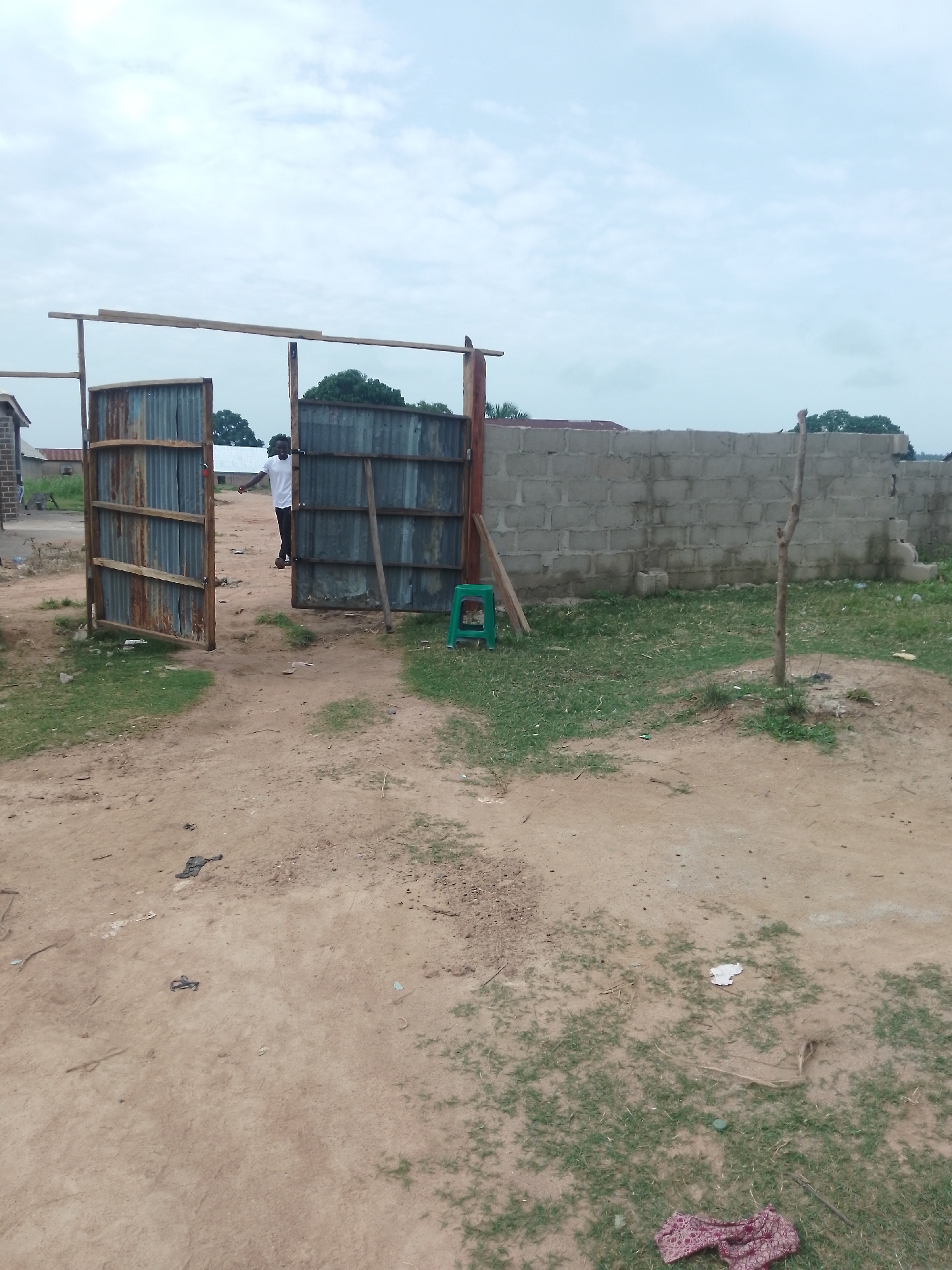 The image shows an outdoor scene in Serti, Nigeria, featuring an unfinished corrugated metal gate supported by wooden posts. A young Black man wearing a white t-shirt and dark trousers stands near the open gate, looking towards the camera. The gate is in front of a wall constructed from concrete blocks. The ground is a mixture of dirt and sparse grass. In the background, there are some buildings and trees. The sky is overcast. To the right of the gate, a green plastic stool is visible. The overall impression is one of an ongoing construction or development project.