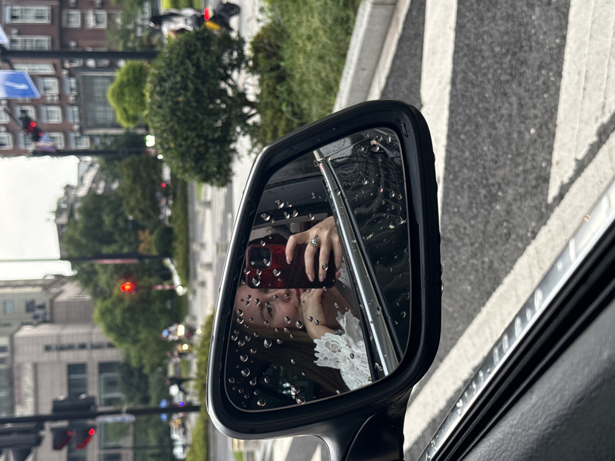 A woman is reflected in a car's side-view mirror, which is covered in raindrops. She is holding a red smartphone up to take a selfie, her face partially obscured by her hand. She wears a white top with lace detailing. The mirror reflects a street scene, including traffic lights, buildings, trees, and a person on a scooter in the background. The scene appears to be in a city, possibly Yiwu, China, given the presence of a red traffic light and the architectural style of the buildings. The road surface is wet, indicating it is raining.