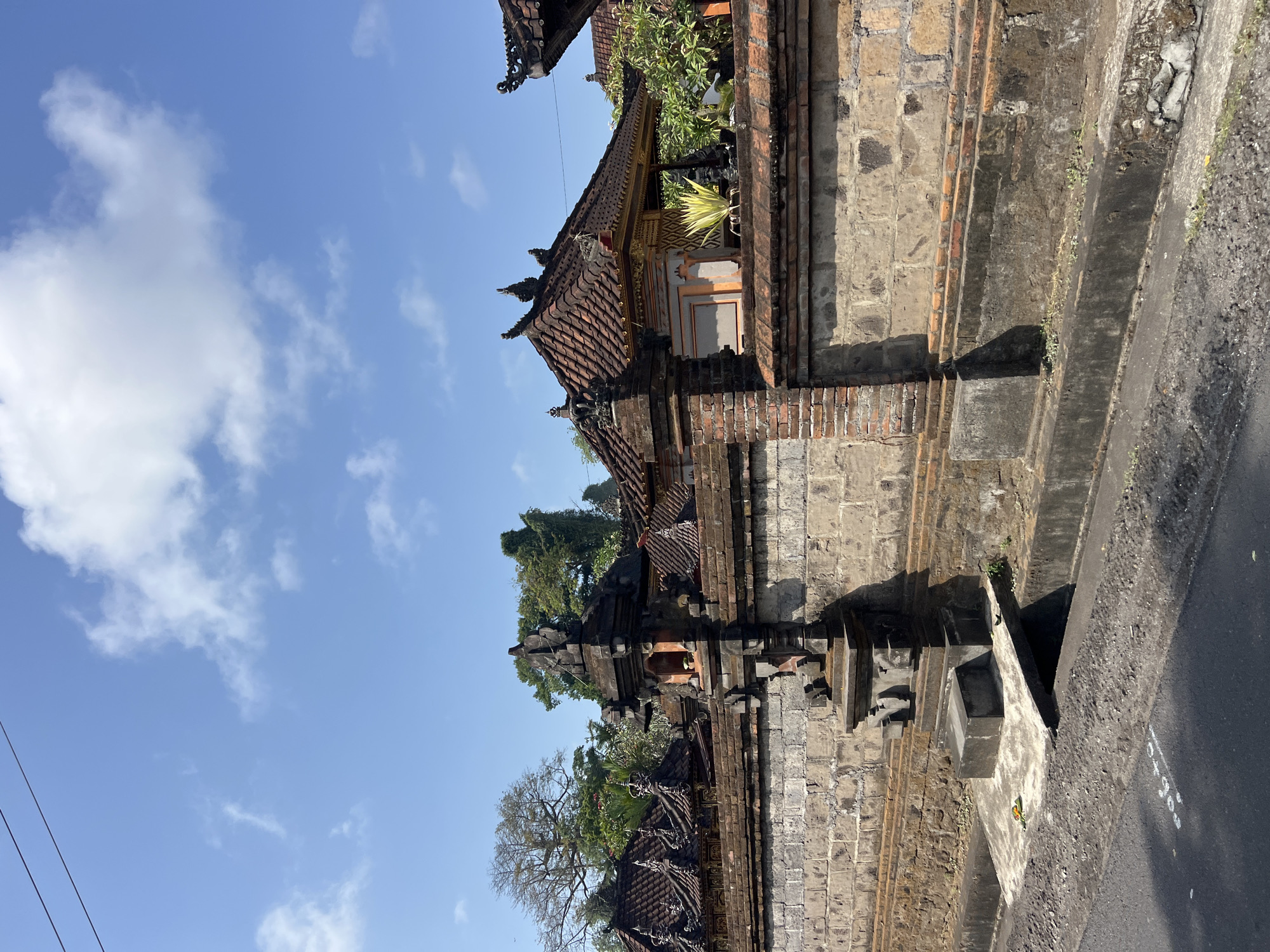 A stone wall with ornate architectural elements lines a street in Sukawati, Indonesia, under a bright blue sky with scattered white clouds. The wall is constructed of large, rough-hewn stone blocks in the lower section and a band of smaller, red brickwork above. Integral to the wall are stone pillars topped with intricate carvings and ornamental rooftops, characteristic of Balinese architecture. Behind the wall, traditional Balinese buildings with multi-tiered, tiled roofs and decorative trim are visible, nestled amongst lush green trees. A narrow concrete drain runs along the base of the wall next to the asphalt road. The scene is brightly lit, suggesting a sunny day.