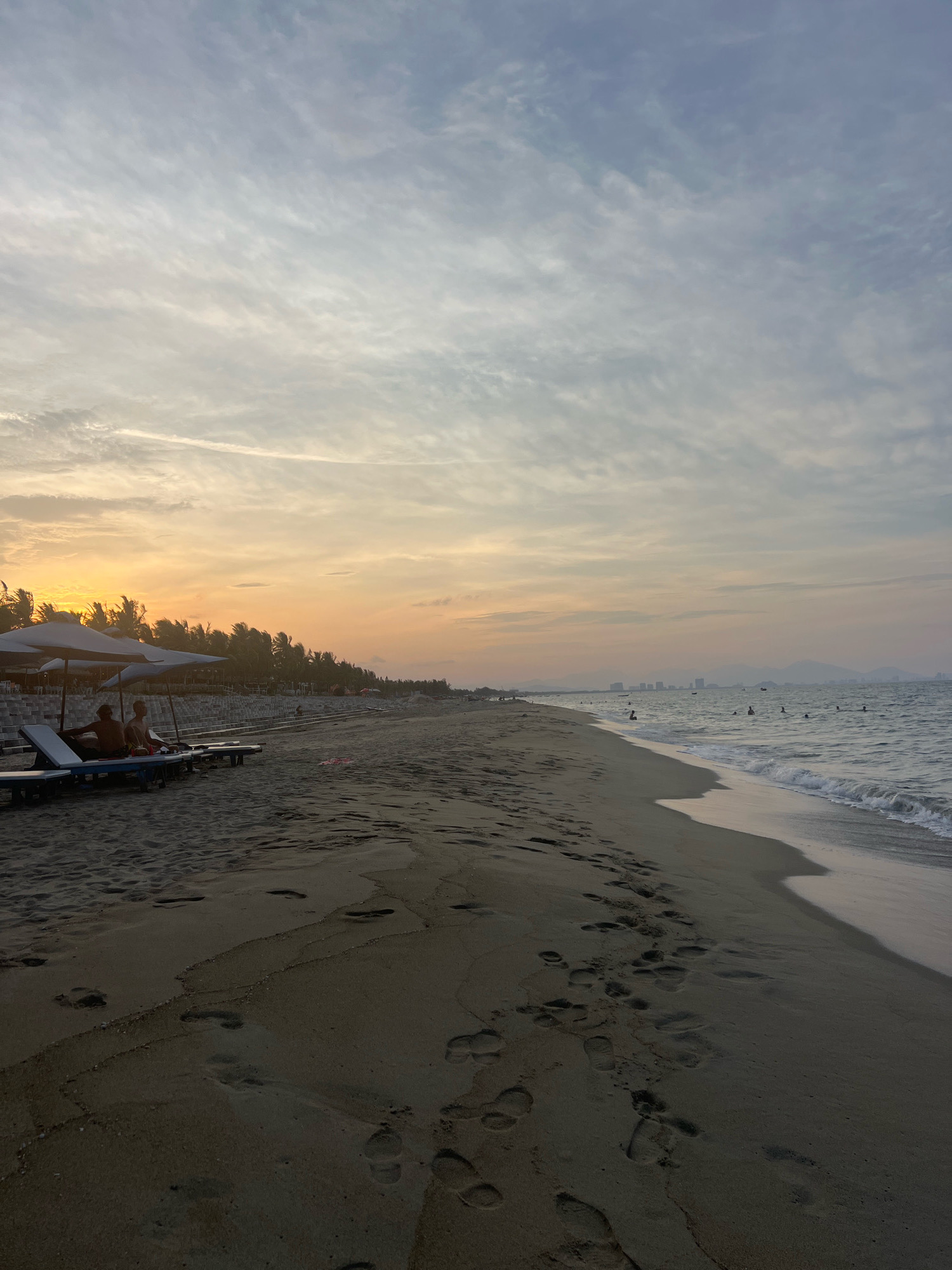 The image depicts a serene sunset scene at An Bang Beach in Hoi An, Vietnam. The sky is a gradient of soft oranges and purples, transitioning into a pale blue, with wispy clouds stretching across. The sandy beach is marked with numerous footprints, indicating recent activity. In the foreground, two lounge chairs are occupied by two individuals, partially shaded by umbrellas, suggesting a moment of relaxation. Further down the beach, a line of palm trees forms a backdrop, with some structures visible behind them. The ocean is calm, with gentle waves lapping at the shore. In the distance, a city skyline can be seen on the horizon, hinting at the proximity of urban development to this coastal paradise. Several people are visible in the water, enjoying a swim as the day concludes.