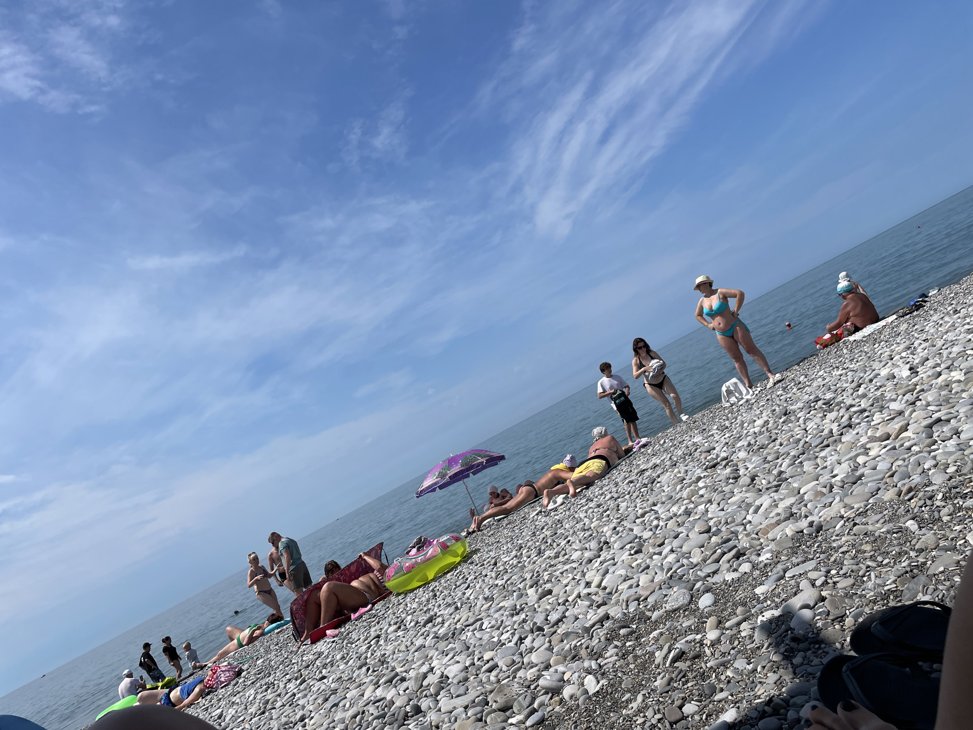 A tilted, eye-level, medium shot captures a sun-drenched, pebble-strewn beach with a group of people sunbathing near the water under a bright, partly cloudy blue sky. The camera angle is set to a slight tilt, adding a dynamic feel to the scene. The majority of the frame is occupied by the beach and the sea, with the sky taking up the upper right portion of the image.
Several individuals are scattered across the beach, some lying down, others sitting, and a few are standing. They are dressed in beach attire, including bikinis, swim trunks, and t-shirts, suggesting a warm-weather outing. One woman in a blue bikini and a straw hat is standing with her hands on her hips, facing away from the camera. Another person in a dark swimsuit is partially visible in the foreground, with their legs and feet in view.
Objects visible on the beach include a purple umbrella, a yellow and pink inflatable ring, and a towel. The pebbles on the beach are varied in size and color, creating a textured surface. The sea is calm and appears to be a deep blue, with a gentle horizon line. The lighting suggests it is daytime, with the sun casting shadows on the beach. The overall atmosphere is one of leisure and relaxation on a summer day.
