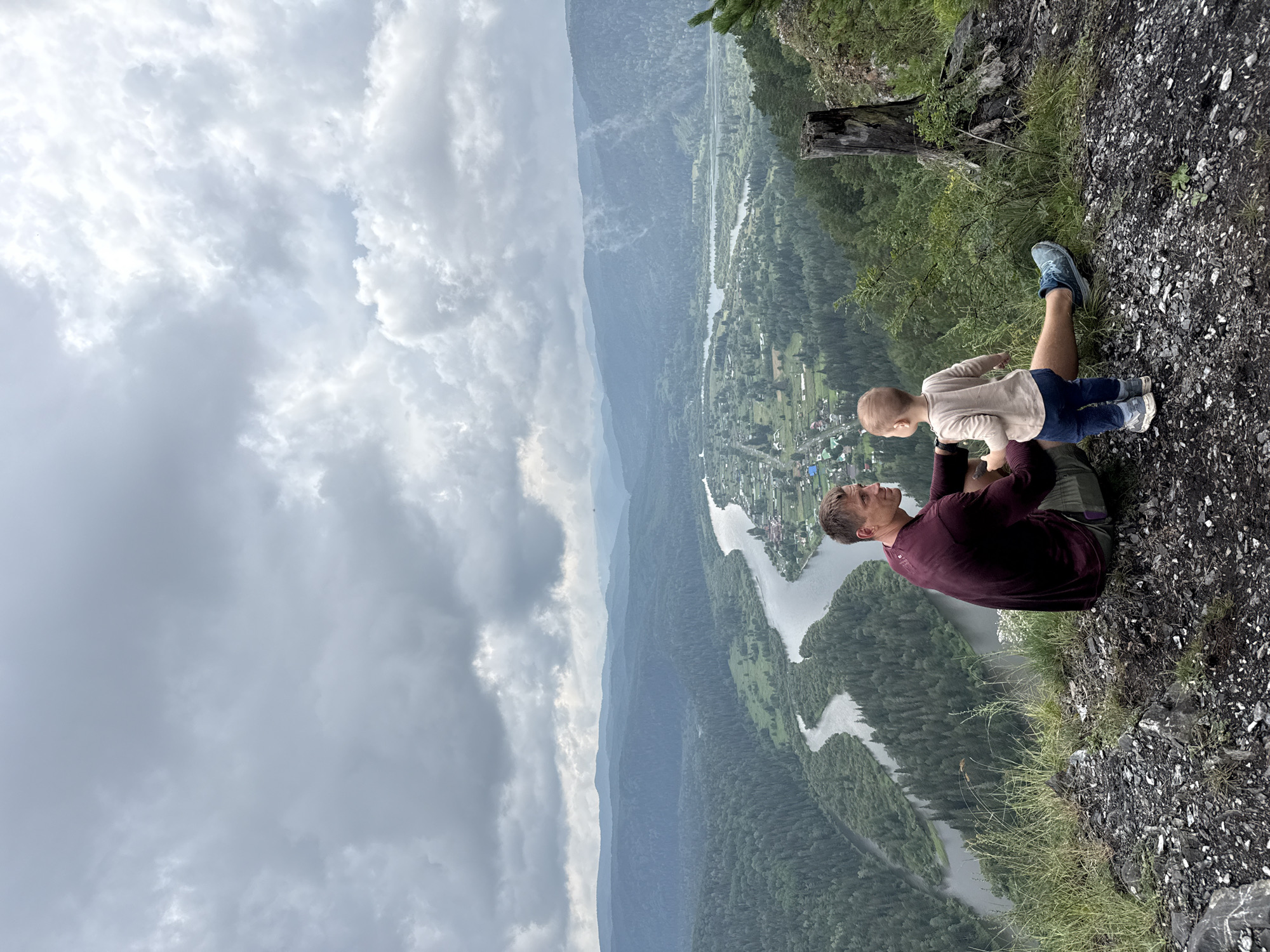 In the city of Urman, Russia, a father and his young child are seated on a rocky ledge overlooking a vast, verdant valley. The father, wearing a maroon long-sleeved shirt and grey shorts, is looking to his right, his gaze directed at the child. The child, dressed in a light beige long-sleeved shirt and dark blue pants, stands facing the father, with their hands extended towards him, seemingly in conversation.
The landscape stretches out before them, a dramatic panorama of rolling hills covered in dense green forest. A wide, winding river snakes through the valley, its waters reflecting the overcast sky. Small villages, with houses nestled among the trees, are visible along the riverbanks. In the foreground, on the right side of the image, a weathered tree stump stands as a silent sentinel on the edge of the precipice. The sky above is a canvas of heavy grey clouds, suggesting the possibility of rain, yet the scene is imbued with a sense of peaceful contemplation.