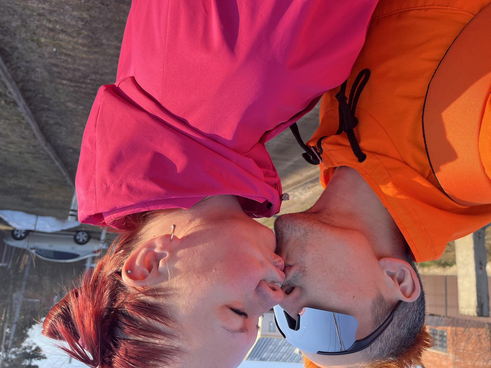 The image shows a couple kissing. The person on the left has red hair and is wearing a pink jacket. The person on the right has short hair, is wearing sunglasses and an orange jacket. The background includes a blurred cityscape with a car visible. The image location may be the city of Chaikovsky, Russia.
