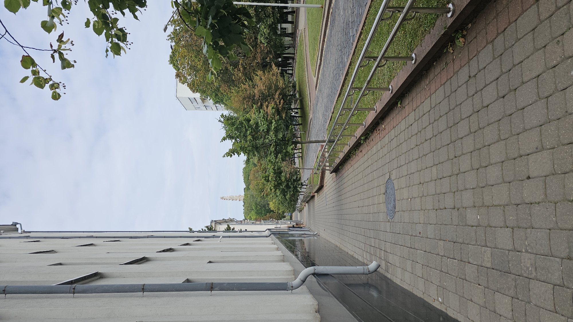 This is a vertical, eye-level, medium shot of a sidewalk next to a building and a street lined with trees. The building is on the left side of the frame and is white with dark grey trim along the bottom. A downspout runs vertically along the building. The sidewalk is made of grey pavers and is empty of people. A manhole cover is visible on the sidewalk. To the right of the sidewalk is a low metal railing. Beyond the railing is a street paved with cobblestones. Trees line the street on the right side of the frame, some with green leaves and others with autumn foliage. In the distance, a tall, light-colored structure resembling a tower or monument is visible. Buildings are also visible in the background among the trees. The sky is overcast with scattered clouds. The scene appears to be a quiet urban street in Minsk, Belarus, likely on a cloudy day in late summer or early autumn.