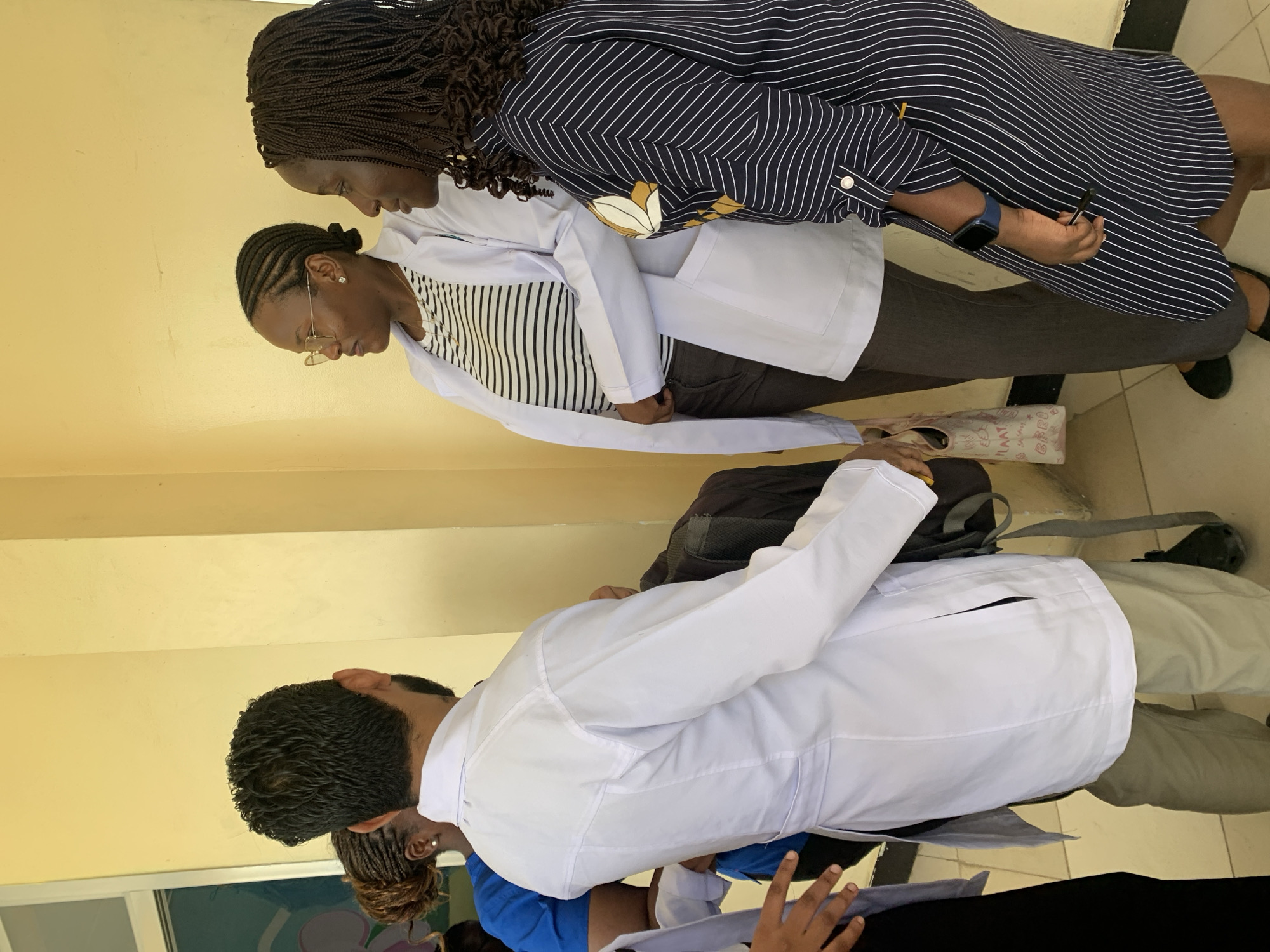 The image captures a group of professionals engaged in an indoor setting, likely a medical or educational facility in Dar es-Salaam, Tanzania. The light yellow walls suggest a well-lit interior.
In the foreground, from left to right, are two women and a man, all appearing attentive. The leftmost woman has cornrows and wears glasses, a vertically striped top, and a white lab coat. She is looking downwards, possibly at an object or document. Next to her, a woman with long braided hair is dressed in a navy blue and white striped dress featuring a subtle floral design. She has a blue smartwatch on her left wrist and holds a pen in her right hand, also directing her gaze downwards.
Behind and to the right, a man is seen from the back, sporting dark curly hair. He wears a white lab coat over a blue scrub top and also appears to be looking down, indicating a shared focal point or discussion among the group. Parts of other individuals, including hands holding potential documents, are visible in the lower right, suggesting more people are present.
Objects visible include white lab coats, a smartwatch, a pen, glasses, a dark backpack worn by the man, and a light-colored tote bag on the floor near the feet of the woman in the striped dress. Partial text reading "ILAAT" and "BBRO" is visible on this tote bag. The overall atmosphere conveys a sense of focus and professionalism.