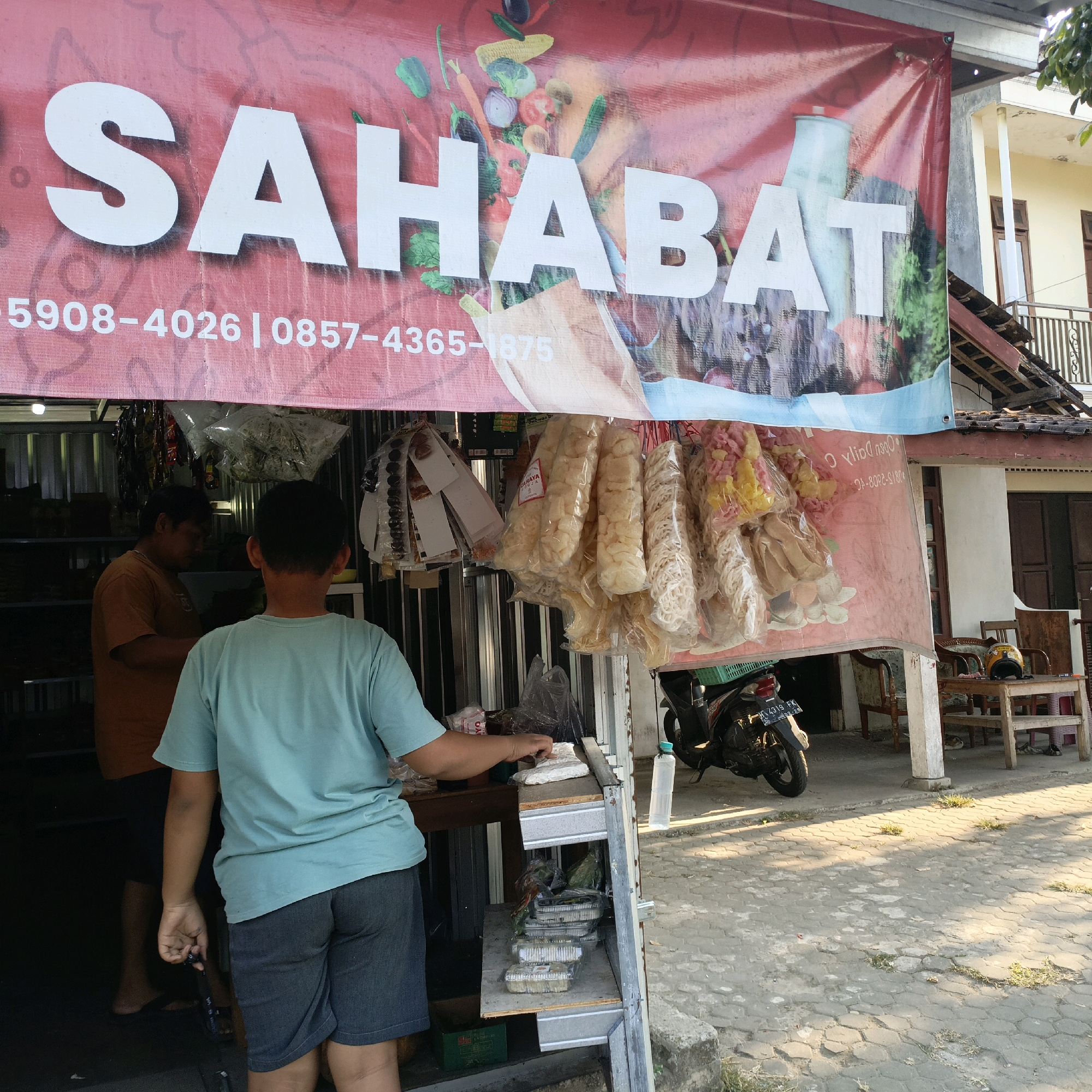 The image shows a street-side shop in Salatiga, Indonesia, with a large red banner proclaiming "SAHABAT" in white lettering. Below the word, phone numbers are visible. Various snacks are displayed hanging in plastic bags in front of the shop. Two people are visible; one, a man in an orange t-shirt, stands inside the shop, while a younger person in a teal t-shirt and denim shorts stands outside, seemingly interacting with the shopkeeper. The shop appears to be an open-air structure with metal siding. In the background, a building with windows and a balcony is visible, along with a parked motorcycle. The ground is paved with interlocking concrete blocks.