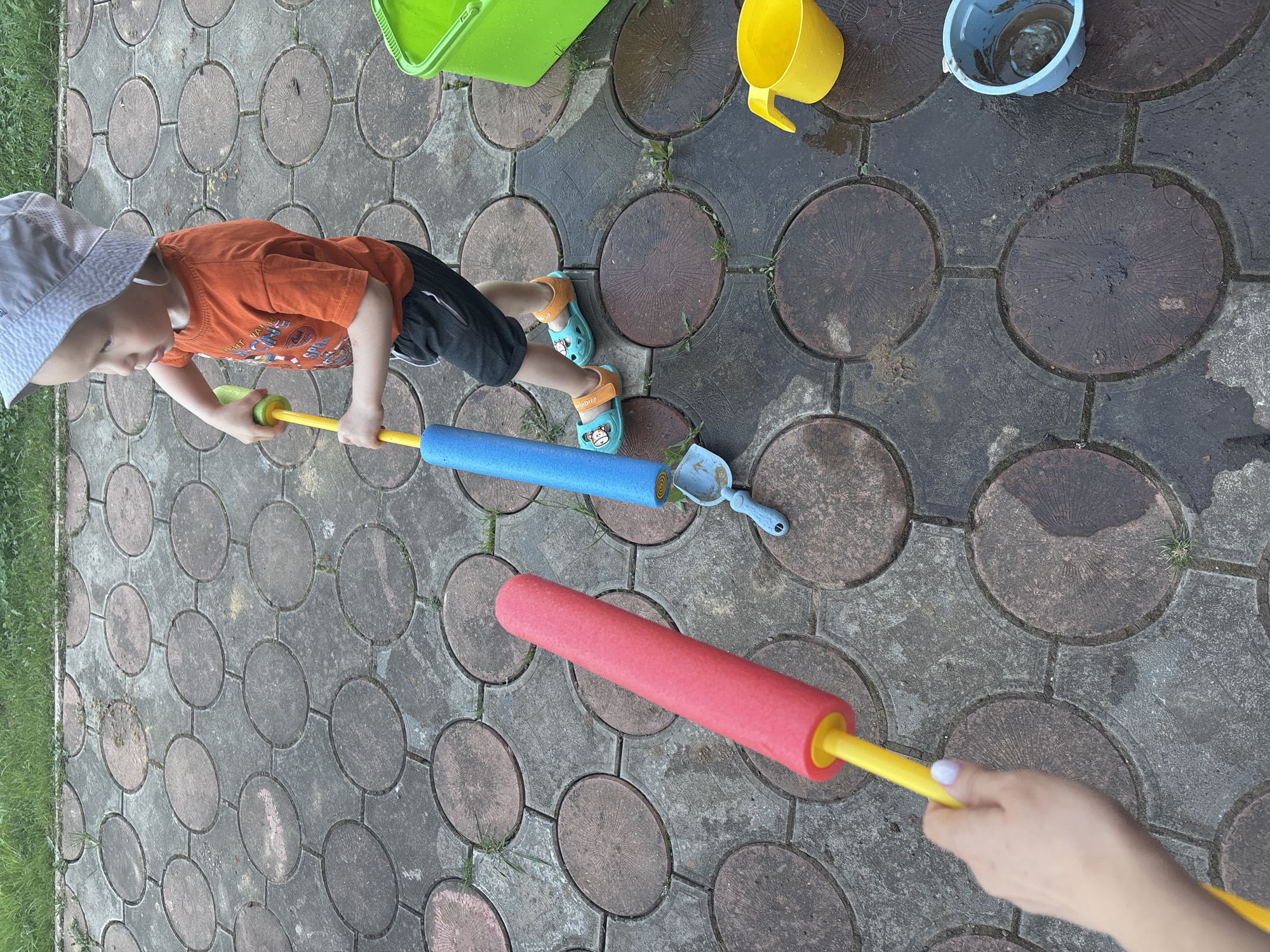 A young child in a white hat, orange shirt, and black shorts stands on a paved surface with circular patterns. They hold a blue foam pool noodle with a yellow handle and a small blue shovel. A red foam pool noodle with a yellow handle is held by a hand in the foreground. Various colored buckets and containers are scattered around. The scene appears to be outdoors, possibly a backyard or patio. The location is Ферма, Russia.